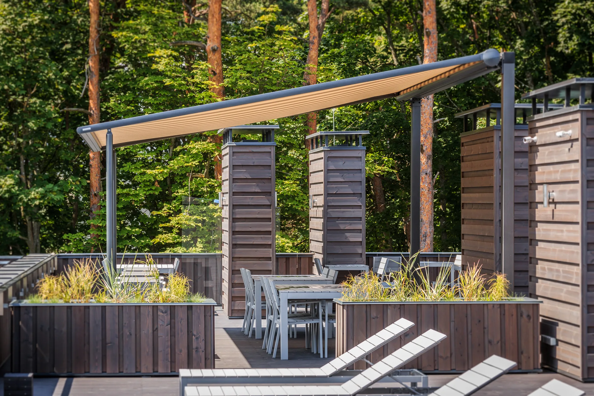 An outdoor rooftop terrace features grey dining tables and loungers beneath a large beige retractable awning, flanked by dark wood privacy columns and planters.