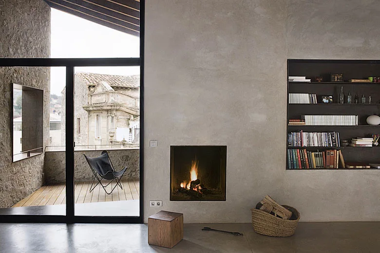A minimalist living room features a plaster wall with an inset fireplace and recessed bookshelf, opening to an outdoor terrace with a butterfly chair.