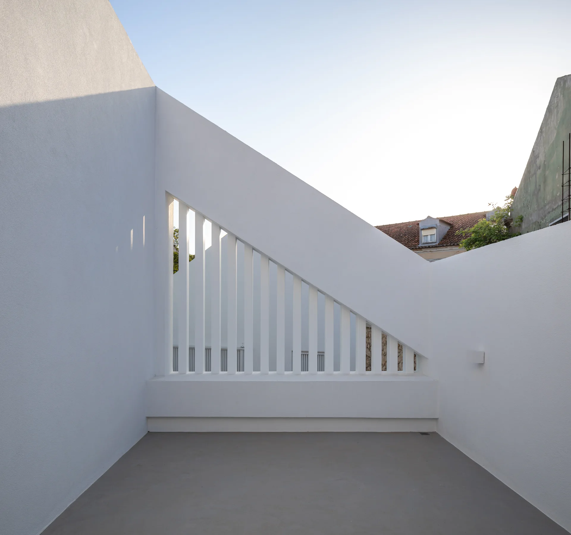 A minimalist white terrace features a concrete floor and a triangular slatted railing integrated into an angular stucco wall.