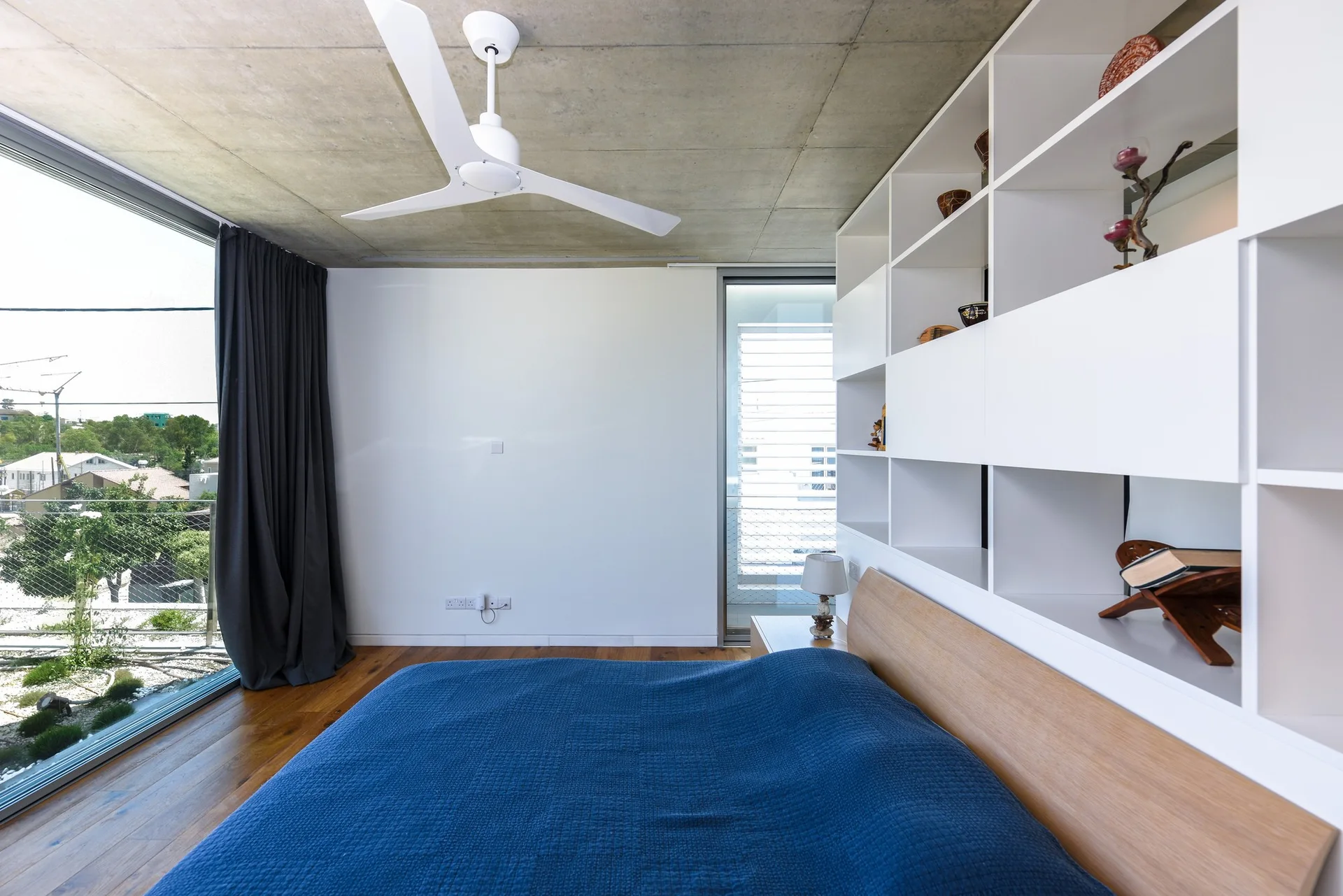 A modern bedroom featuring an exposed concrete ceiling, a wood bed with a blue duvet, and a large white built-in shelving unit.