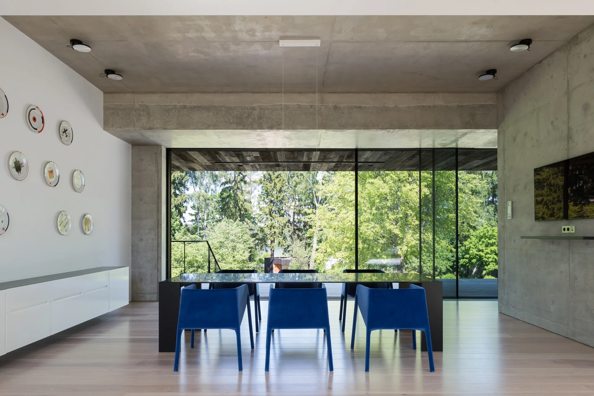 A modern dining room features cobalt blue chairs around a black table beneath a raw concrete ceiling with glass walls overlooking trees.