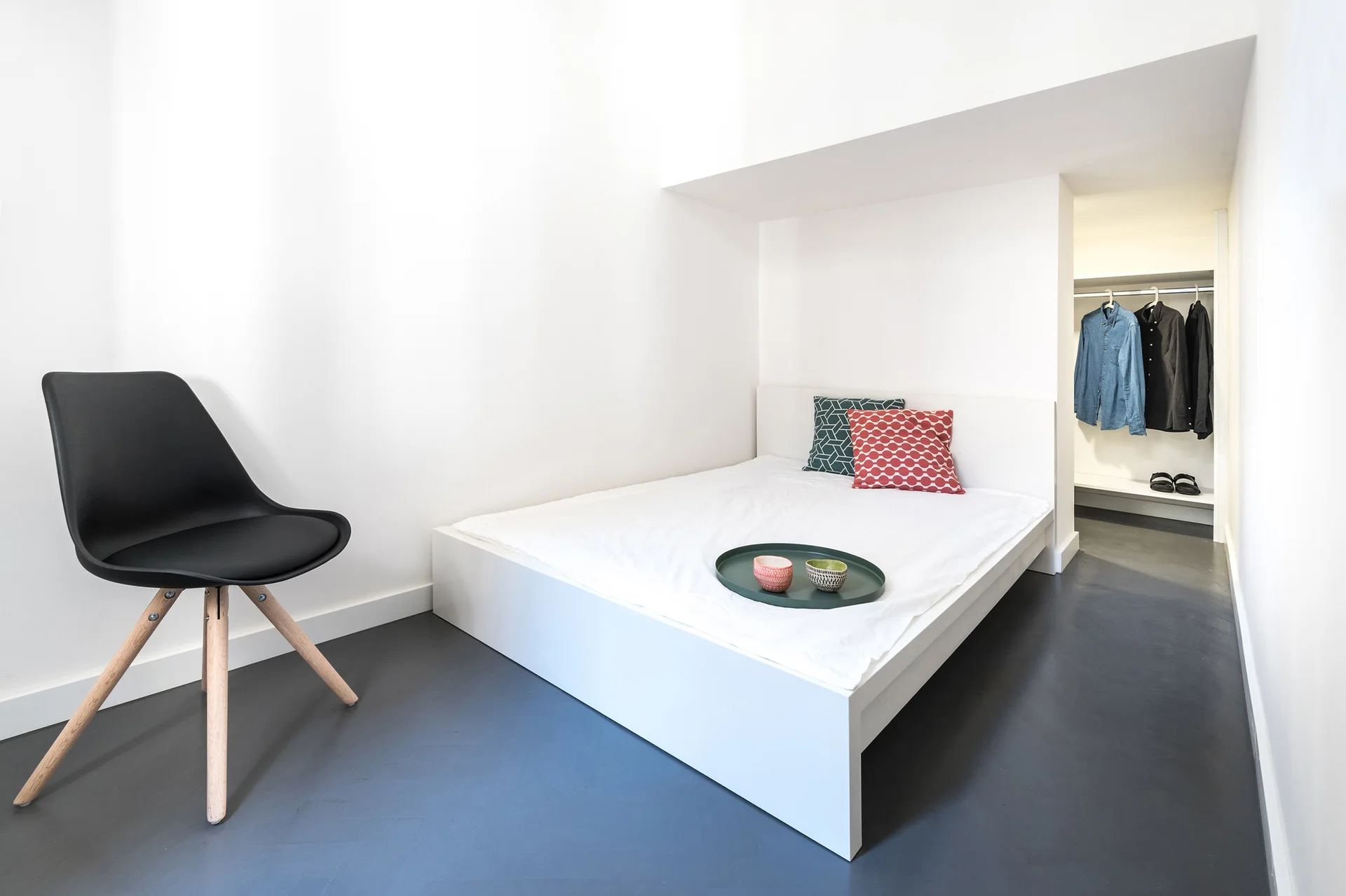 A minimalist white bedroom featuring a platform bed in an alcove, a black molded chair, dark grey flooring, and an open recessed wardrobe.