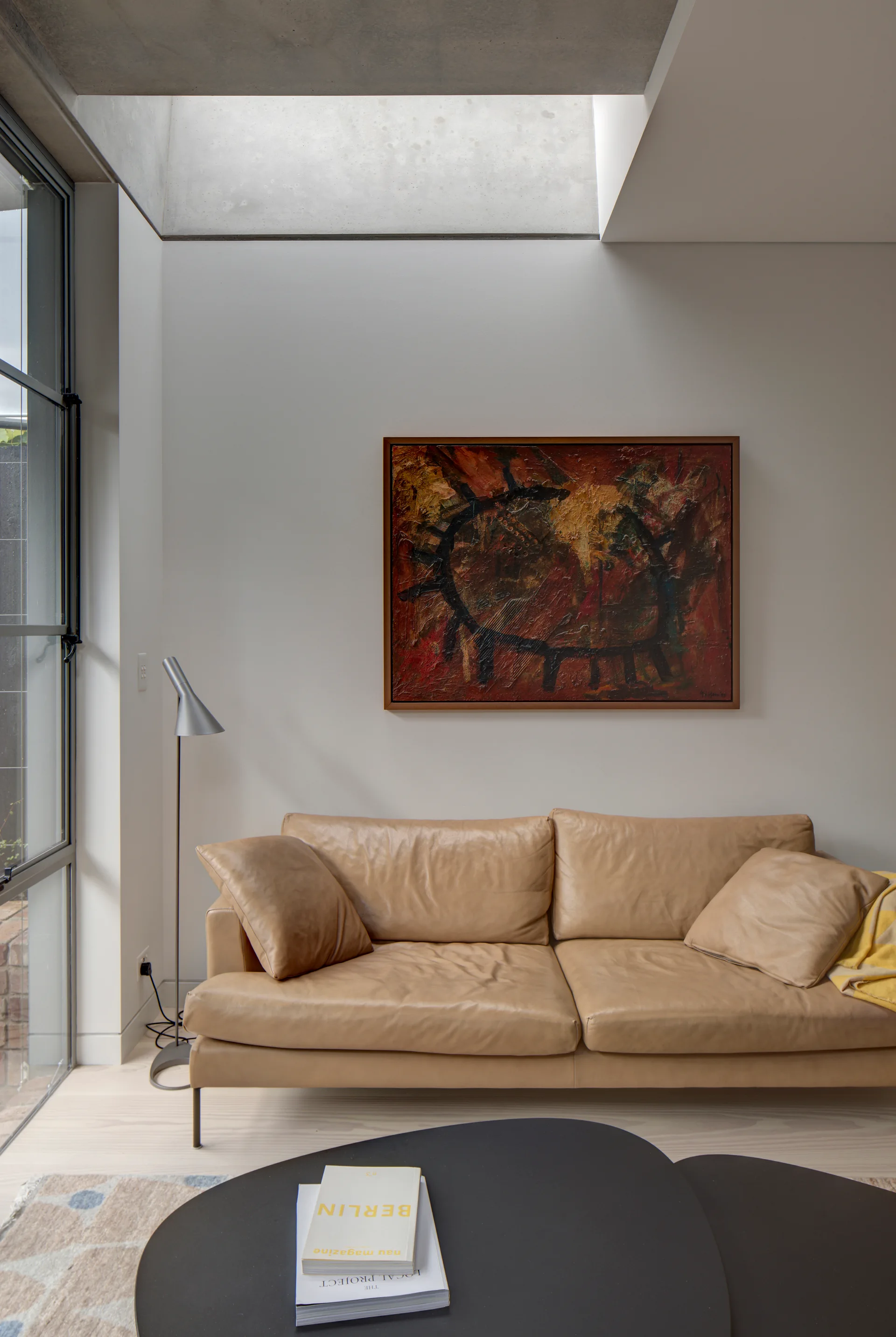 A tan leather sofa sits beneath a framed abstract painting in a bright living room featuring a concrete skylight recess and black coffee table.
