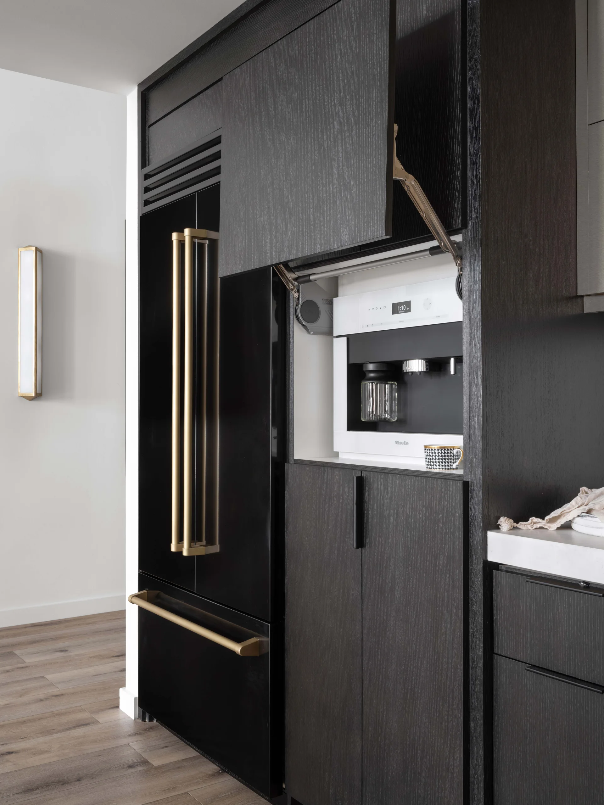 Modern kitchen featuring dark wood grain cabinets, a built-in white coffee machine behind a lift-up door, and a black refrigerator with gold handles.