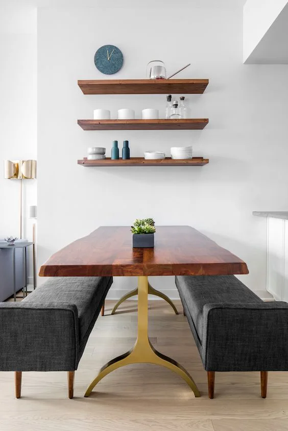 A wooden live-edge dining table with a gold base is flanked by two gray upholstered benches under three floating shelves on a white wall.