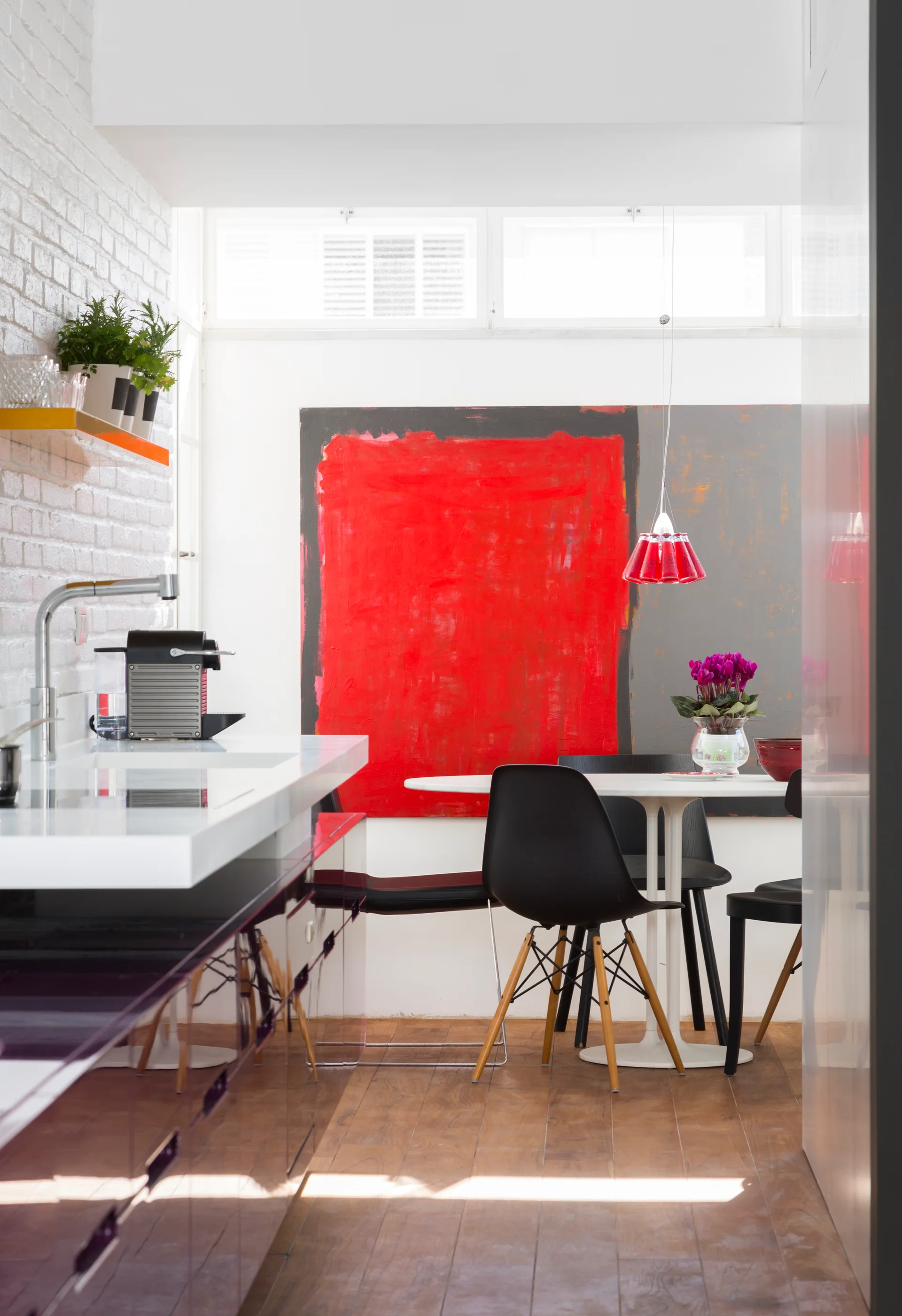 A modern kitchen and dining space featuring white painted brick, glossy purple cabinets, a round dining table with black chairs, and large red artwork.