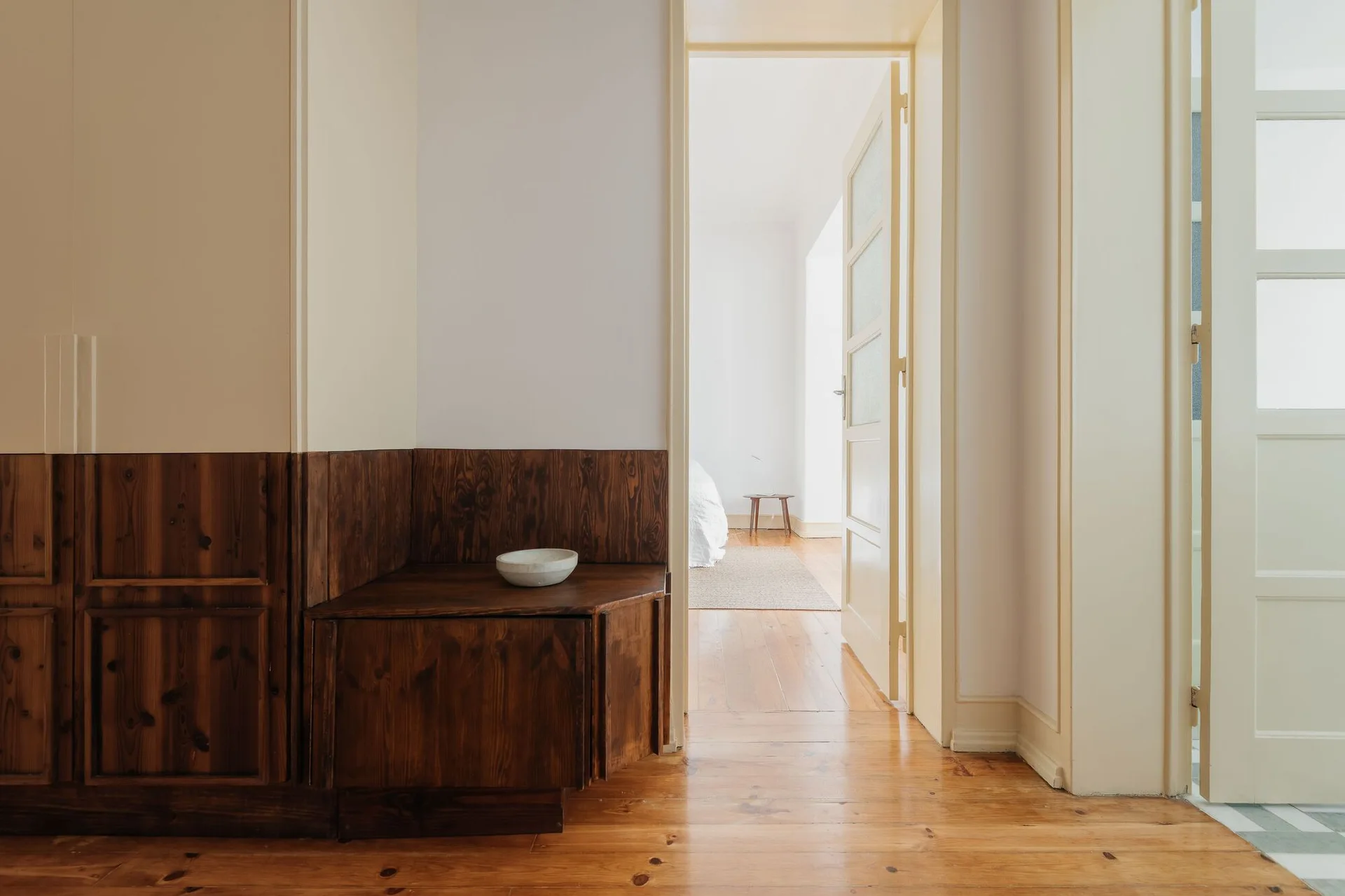 A bright hallway features warm pine floors, white walls with dark stained wood wainscoting, and a built-in corner cabinet holding a ceramic bowl.