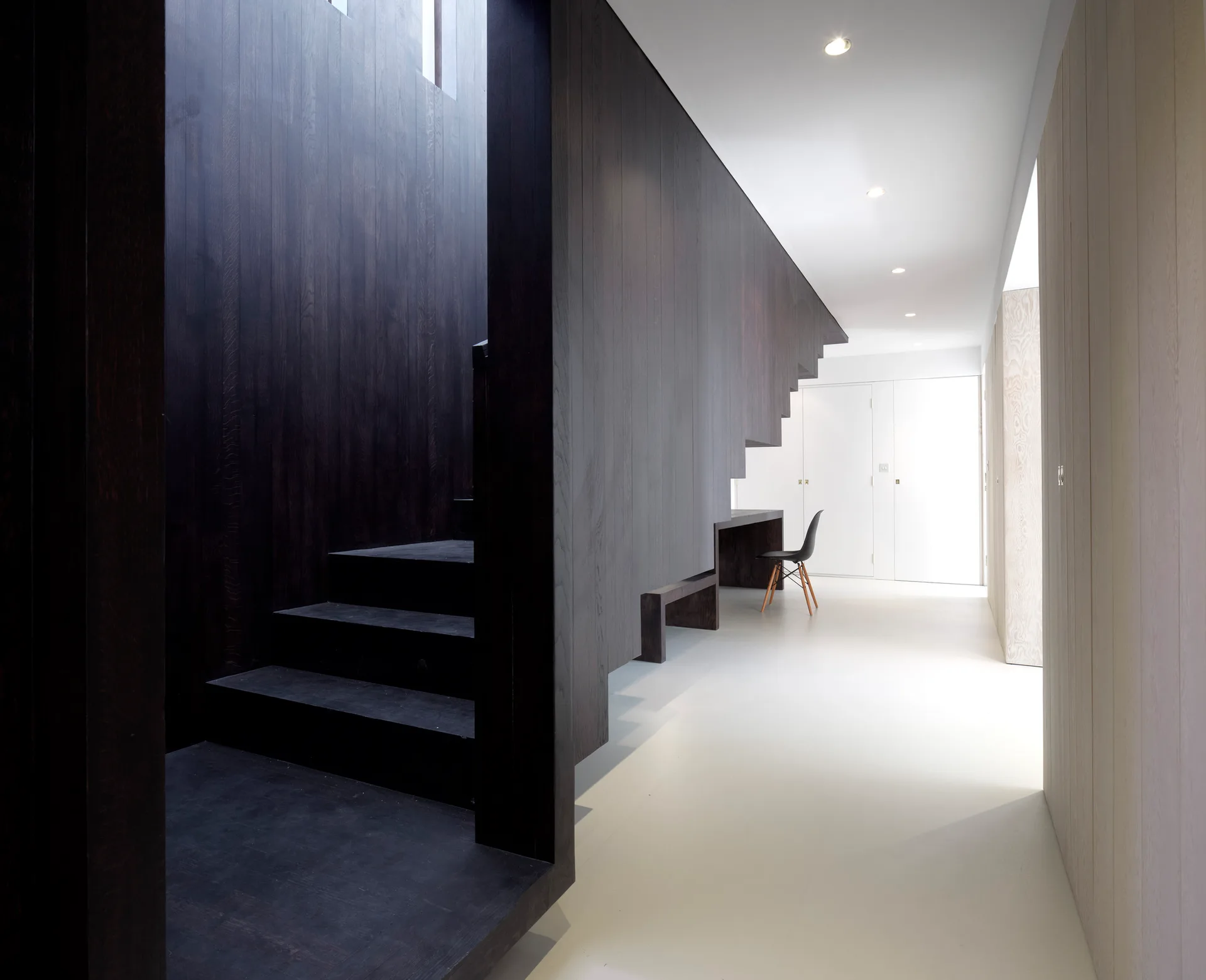 A dark wood paneled staircase with an integrated built-in desk stands in a minimalist white hallway with a black chair.