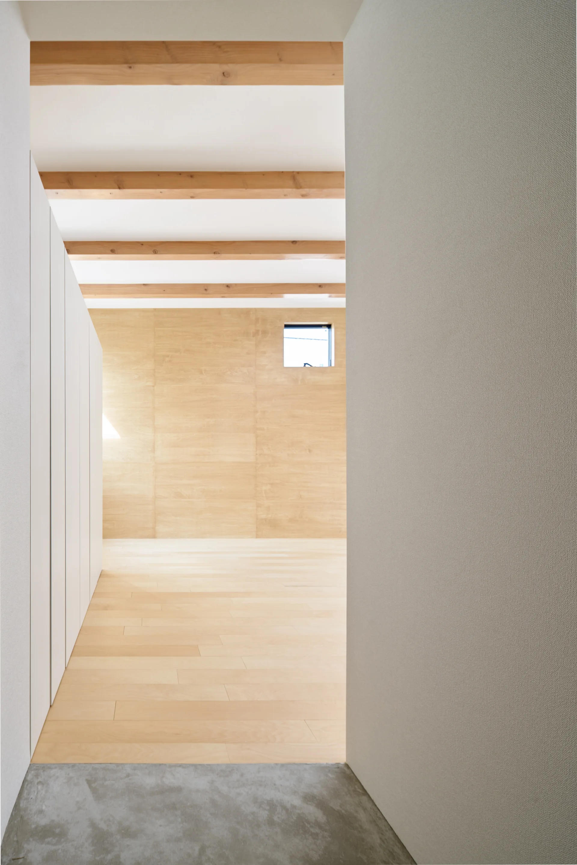 A view from a grey concrete entryway into a minimalist room with light wood flooring, wood-paneled walls, and exposed ceiling beams.