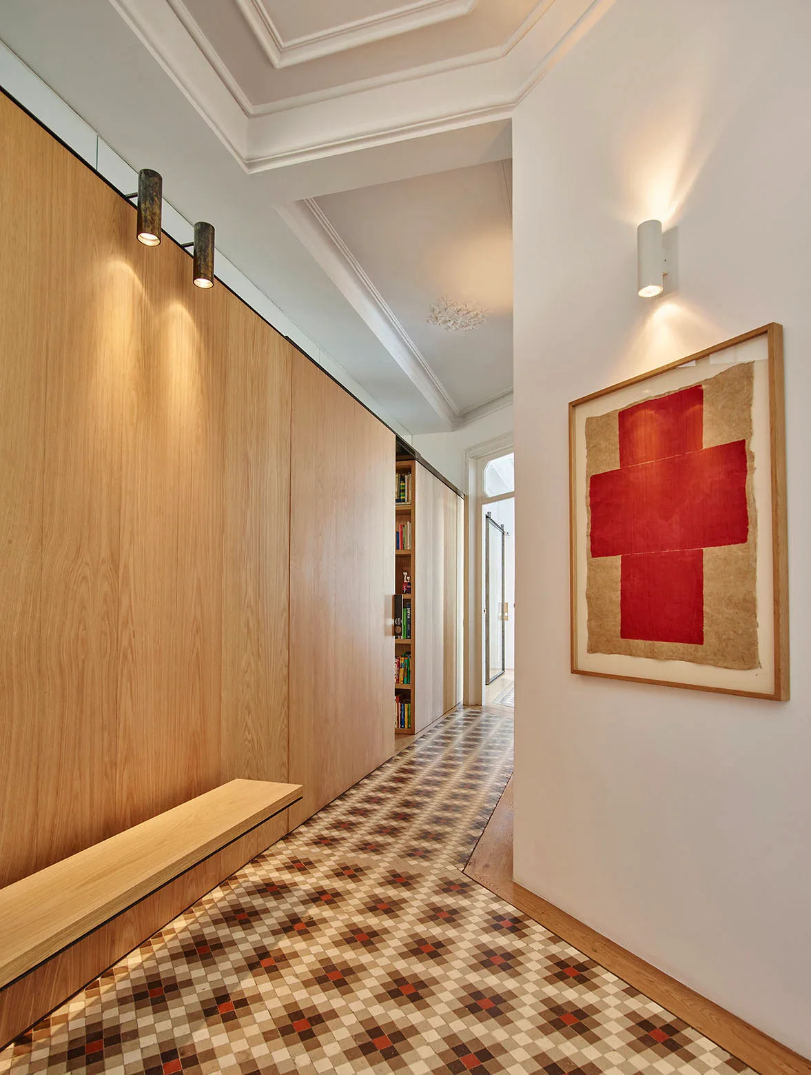 A hallway featuring full-height light wood wall paneling, a patterned checkered tile floor, a floating wooden bench, and a framed red cross artwork.