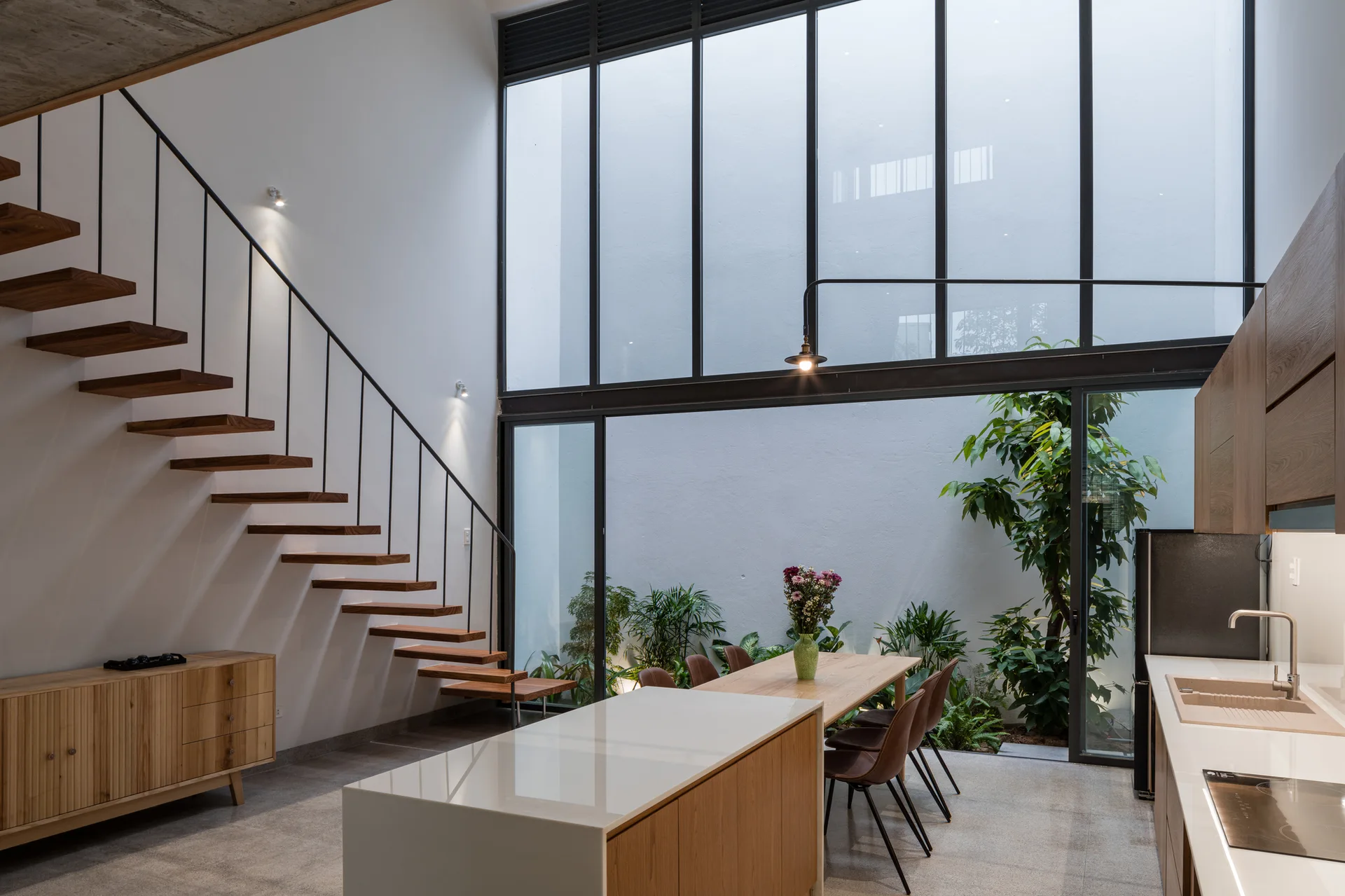 A modern kitchen and dining area with a wood cantilevered staircase and a glass wall looking into a plant-filled courtyard.