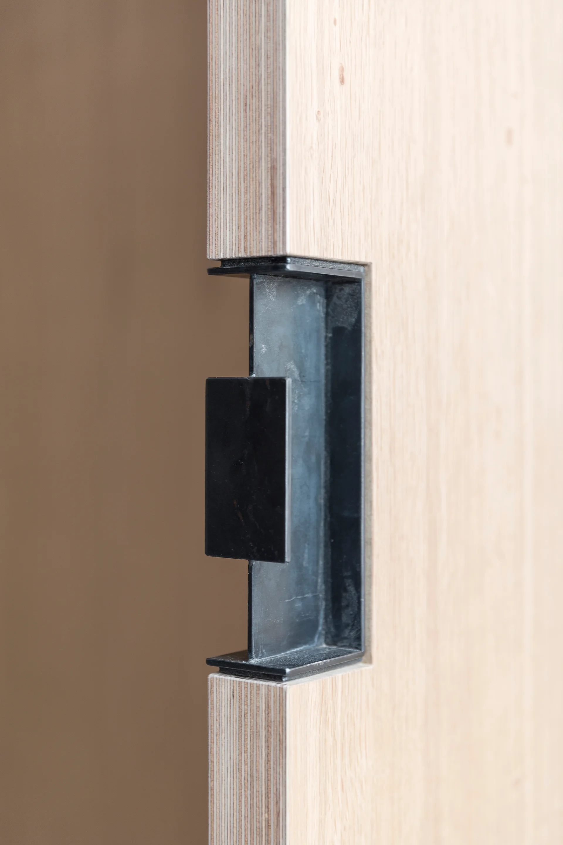 A close-up of a matte black recessed flush pull handle integrated into the layered edge of a light wood plywood door.