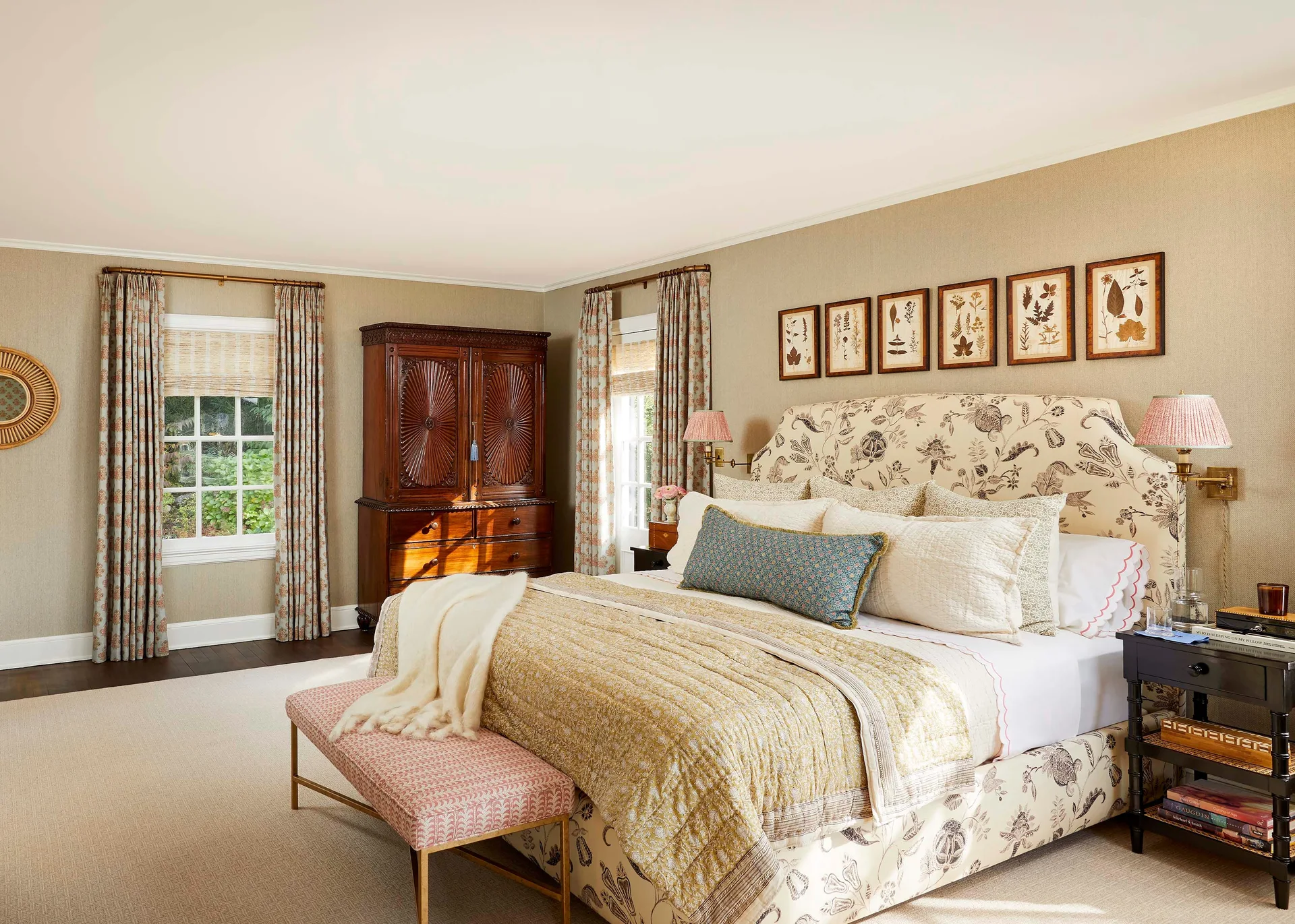 A traditional bedroom features an upholstered floral bed, a series of six framed botanical prints, and a large carved wooden armoire.