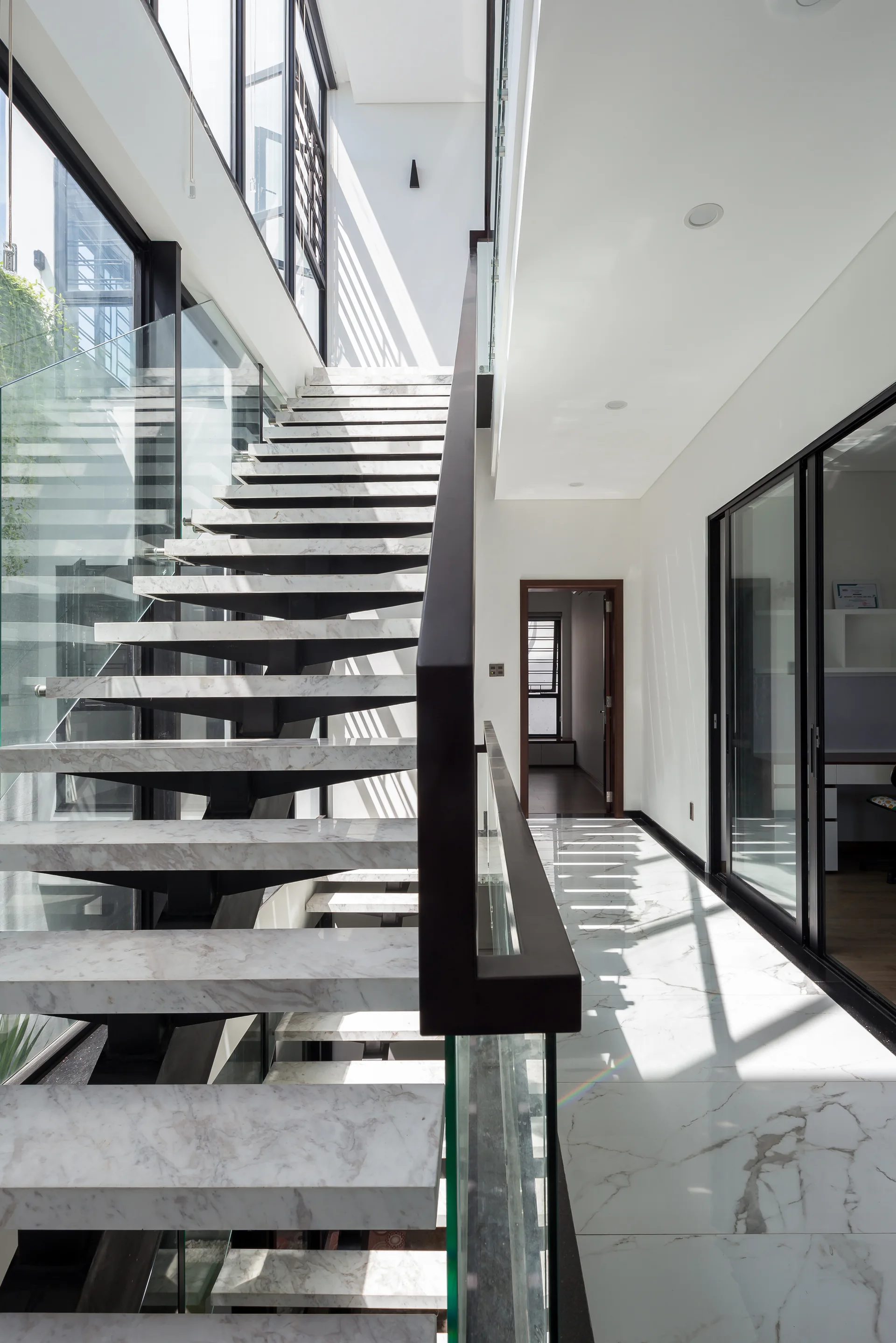 A modern staircase with floating white marble treads and a glass railing stands next to a polished marble hallway floor under bright sunlight.