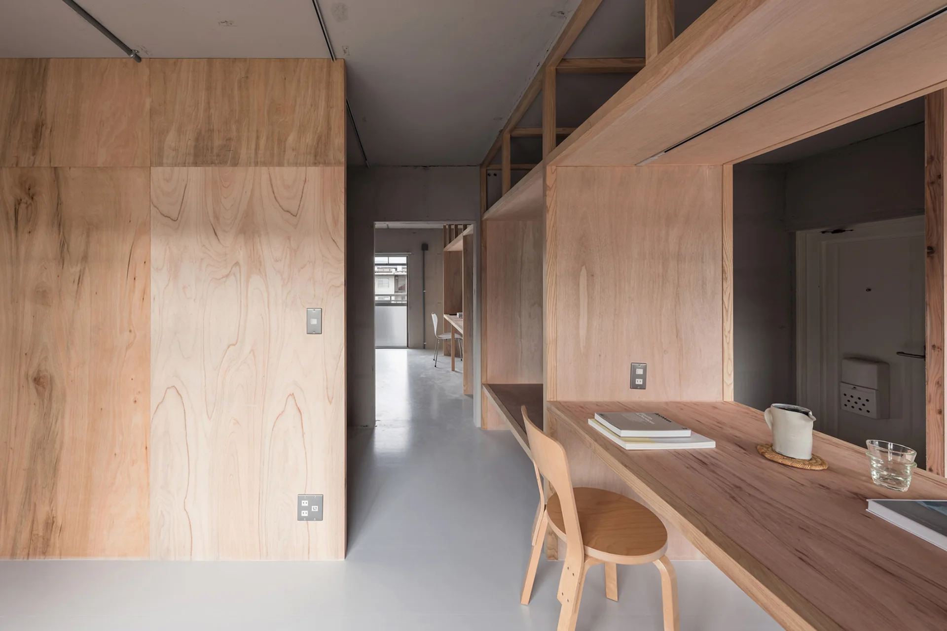 A minimalist study featuring walls clad in light plywood panels, a matching built-in wooden desk with a chair, and a smooth grey floor.