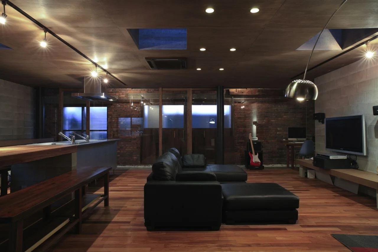 An open-plan living room featuring a dark wood paneled ceiling with skylights, a black leather sectional sofa, and exposed brick walls.