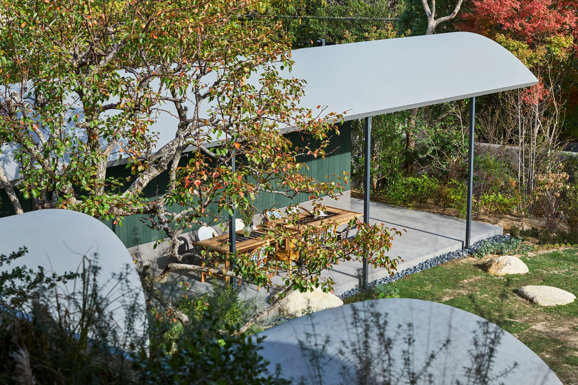 A long wooden dining table and chairs sit on a concrete patio beneath a curved white metal canopy next to a dark green house siding.