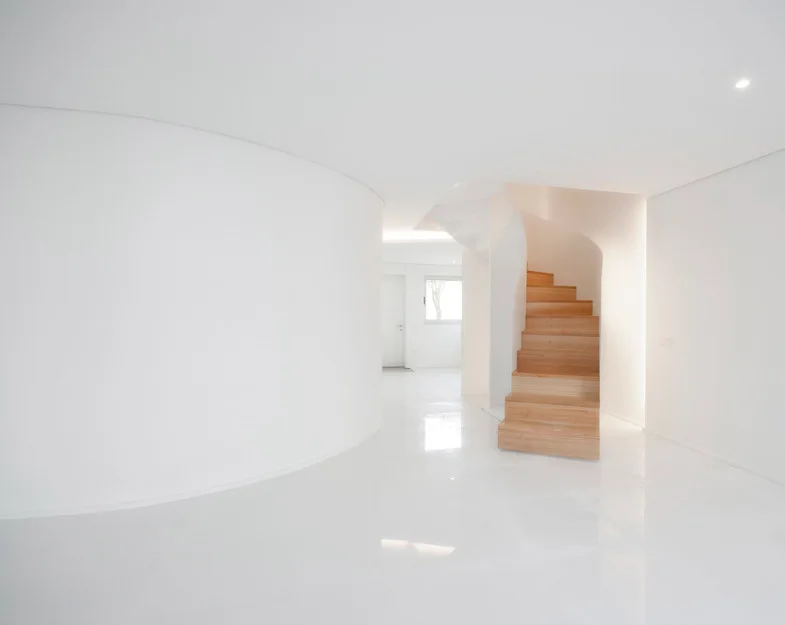 A minimalist white foyer featuring a large curved wall, glossy reflective floors, and a wooden spiral staircase.
