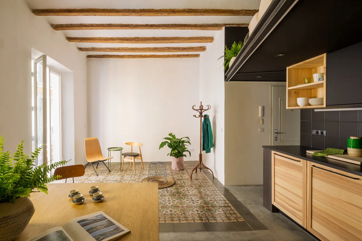 An open-concept room features exposed timber ceiling beams, patterned ceramic floor tiles, and wood and black kitchen cabinetry.