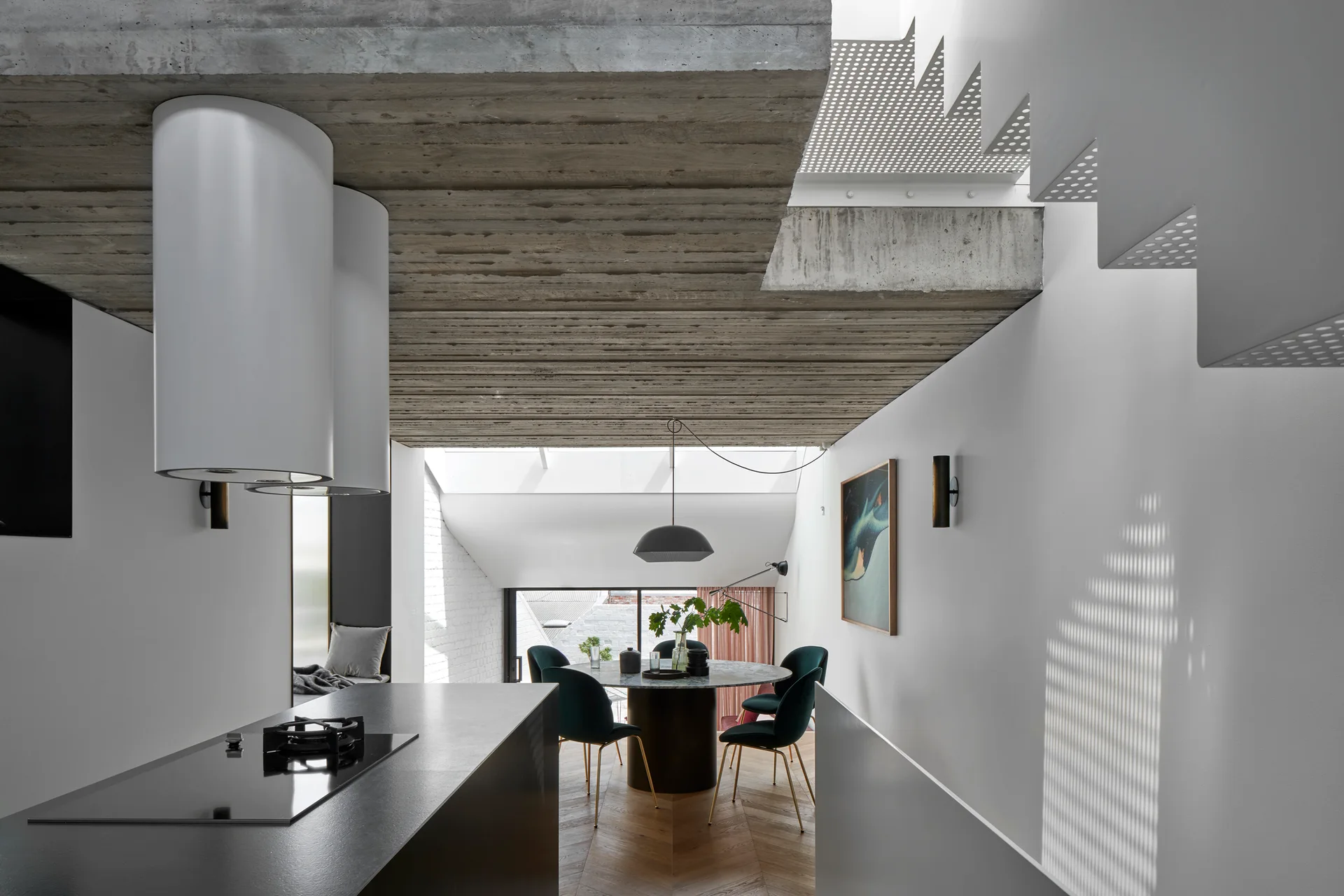 An industrial kitchen with a dark island and white range hoods opens to a dining area beneath an exposed concrete ceiling and skylight.