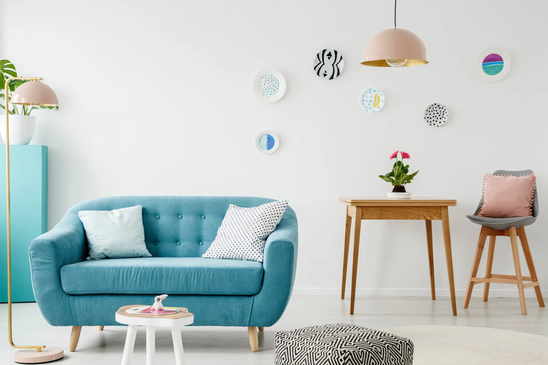A bright living room features a tufted blue sofa, a wooden table, and decorative plates arranged on a white wall.