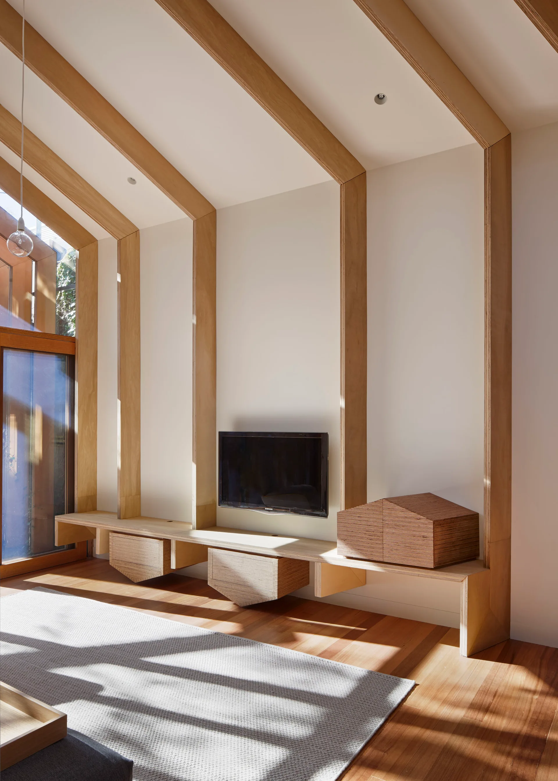 A minimalist living room with white walls, exposed plywood ceiling beams, a wall-mounted TV, and a floating wooden media console over wood floors.