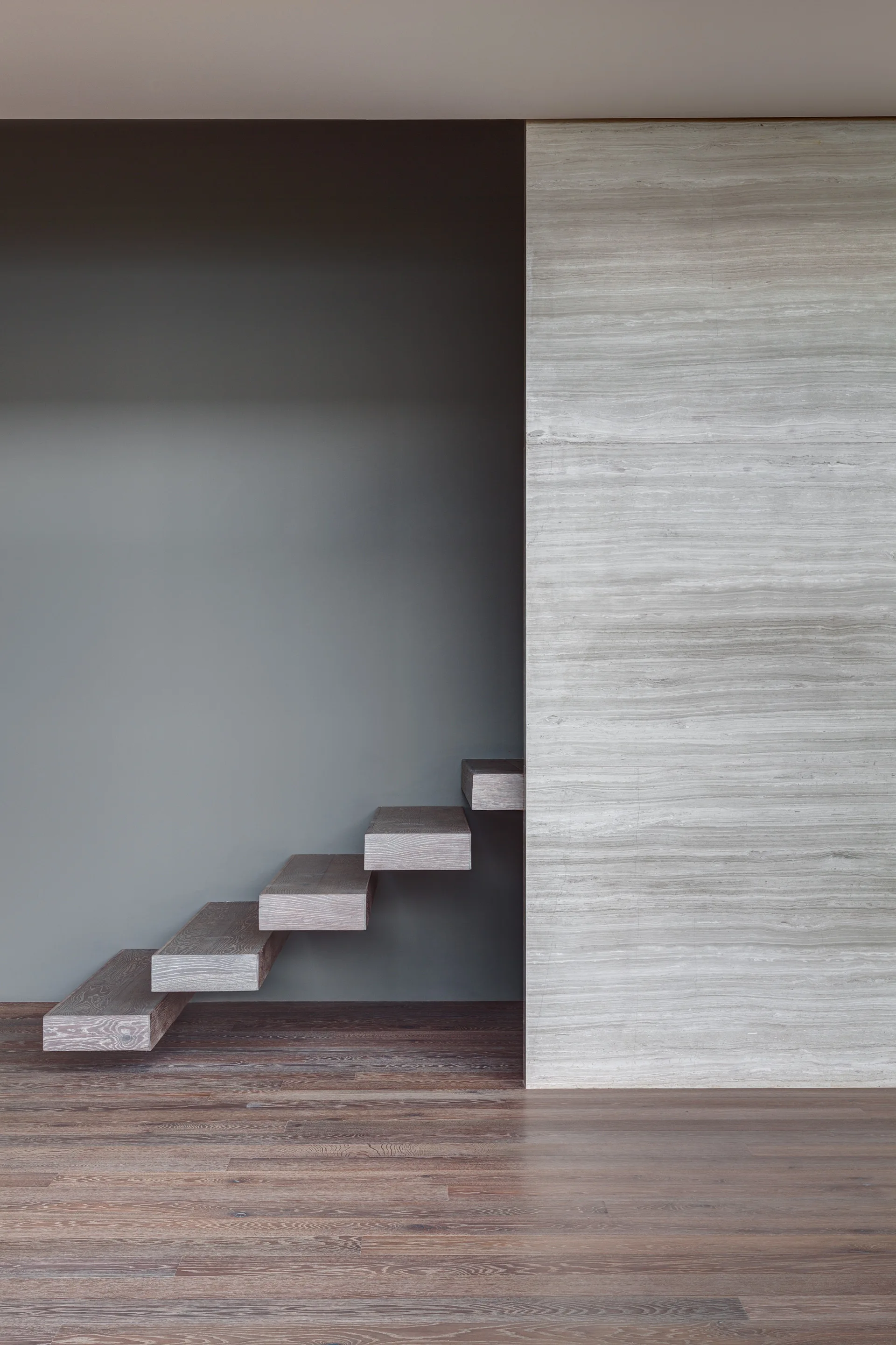 Minimalist floating wood stairs cantilever out from a matte grey wall next to a large veined stone panel and dark wood flooring.