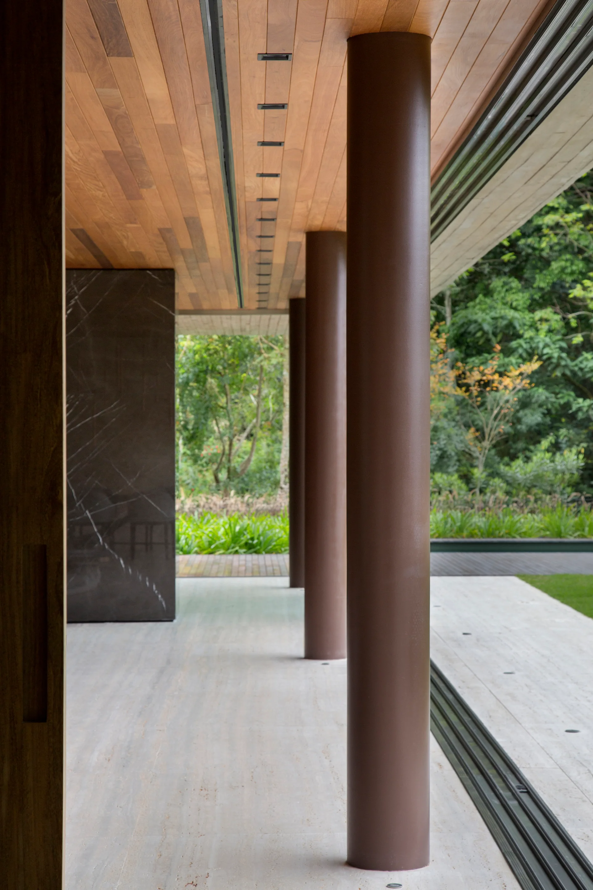 A modern covered patio featuring a warm wood ceiling, light stone tile floors, and a row of cylindrical columns bordering a green lawn.