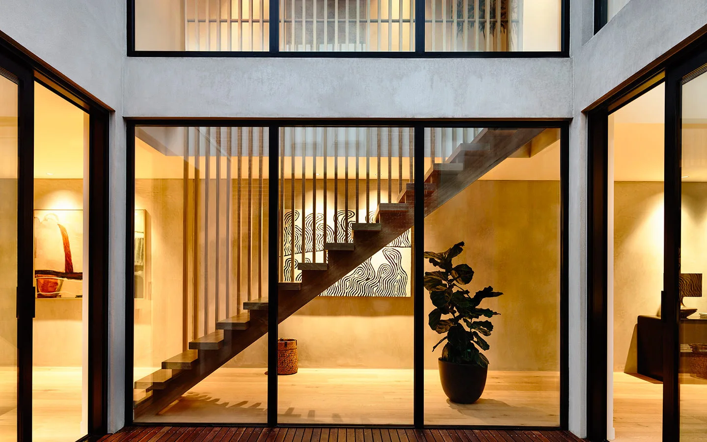 A modern floating wooden staircase with a vertical slat railing is visible through large black-framed glass walls in a warmly lit home.
