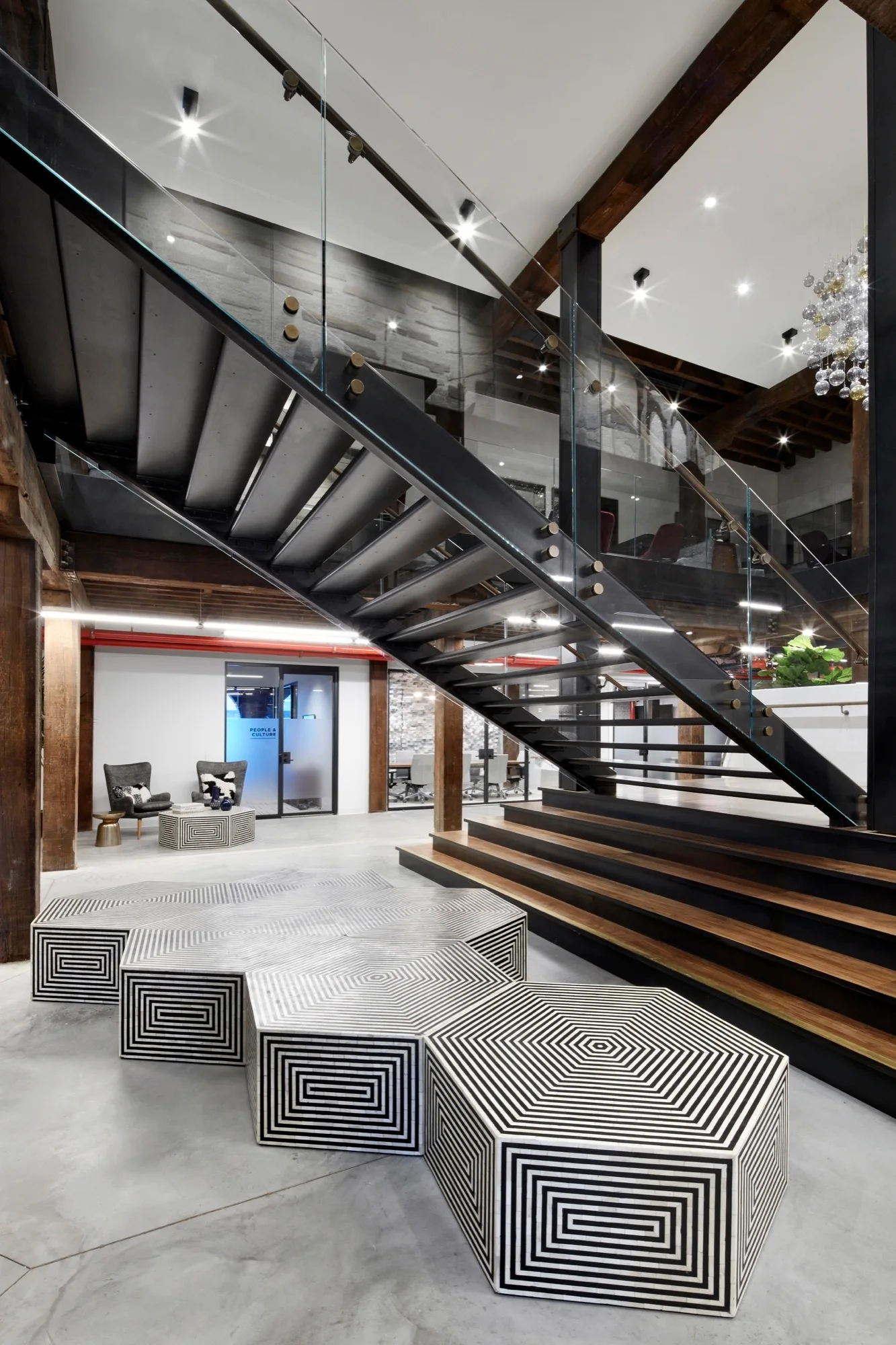 A modern industrial lobby features a metal staircase with glass railings, rustic wood beams, and modular black and white geometric ottomans on a concrete floor.