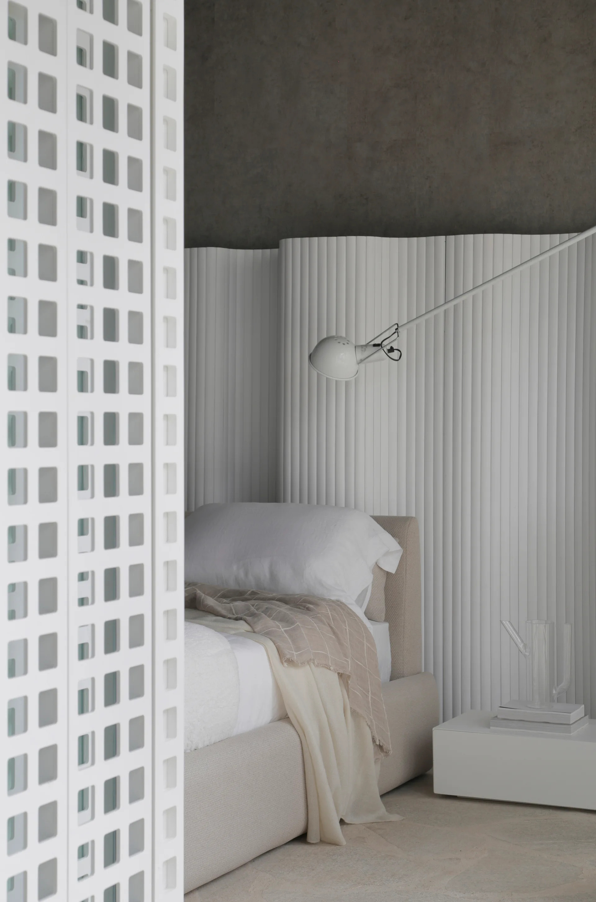A white upholstered bed is placed against a curved fluted wall panel next to an adjustable white reading lamp and a geometric room divider.