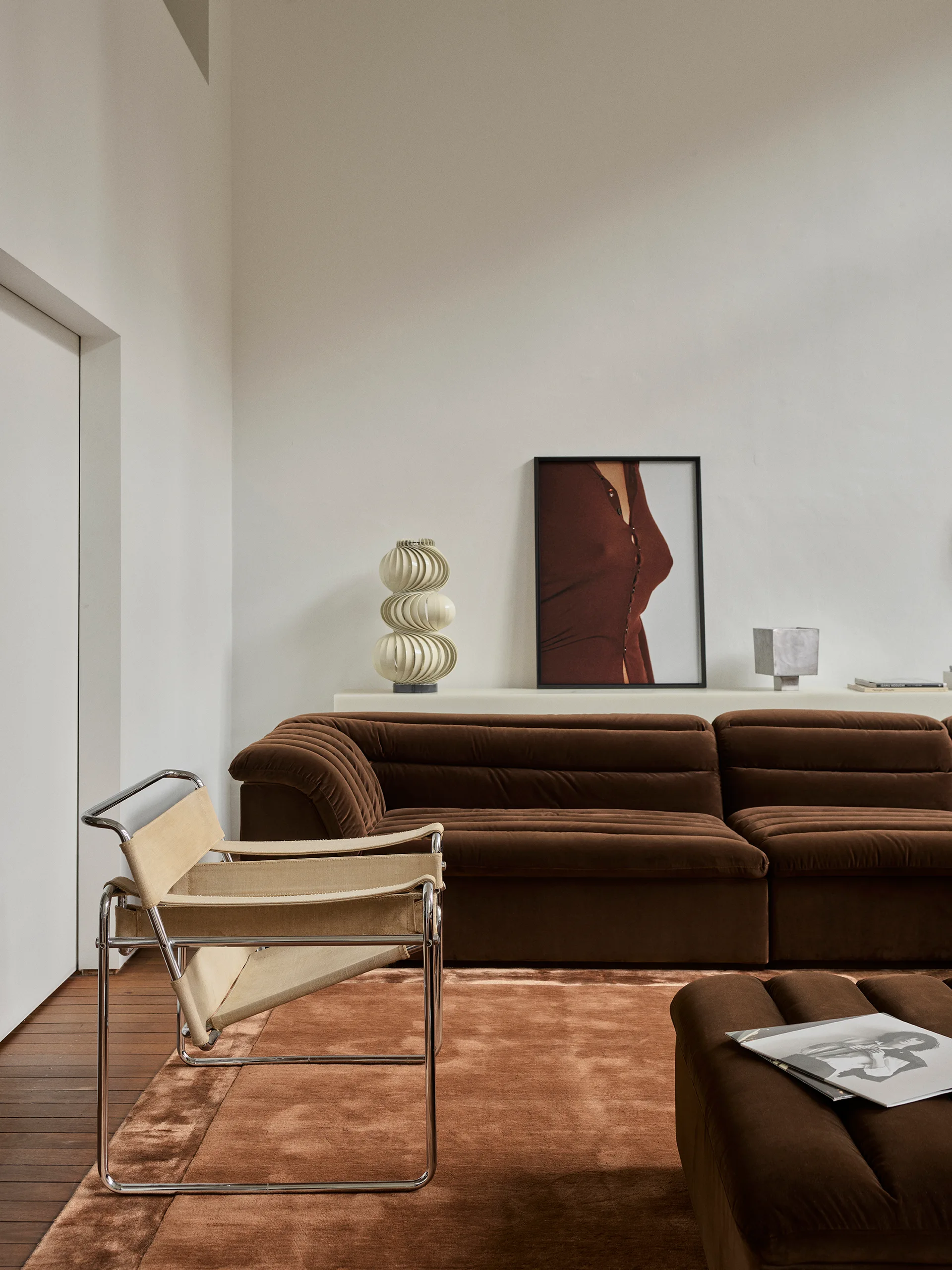A chocolate brown channel-tufted sofa sits next to a tubular chrome-framed canvas chair on a plush brown rug against an off-white wall.