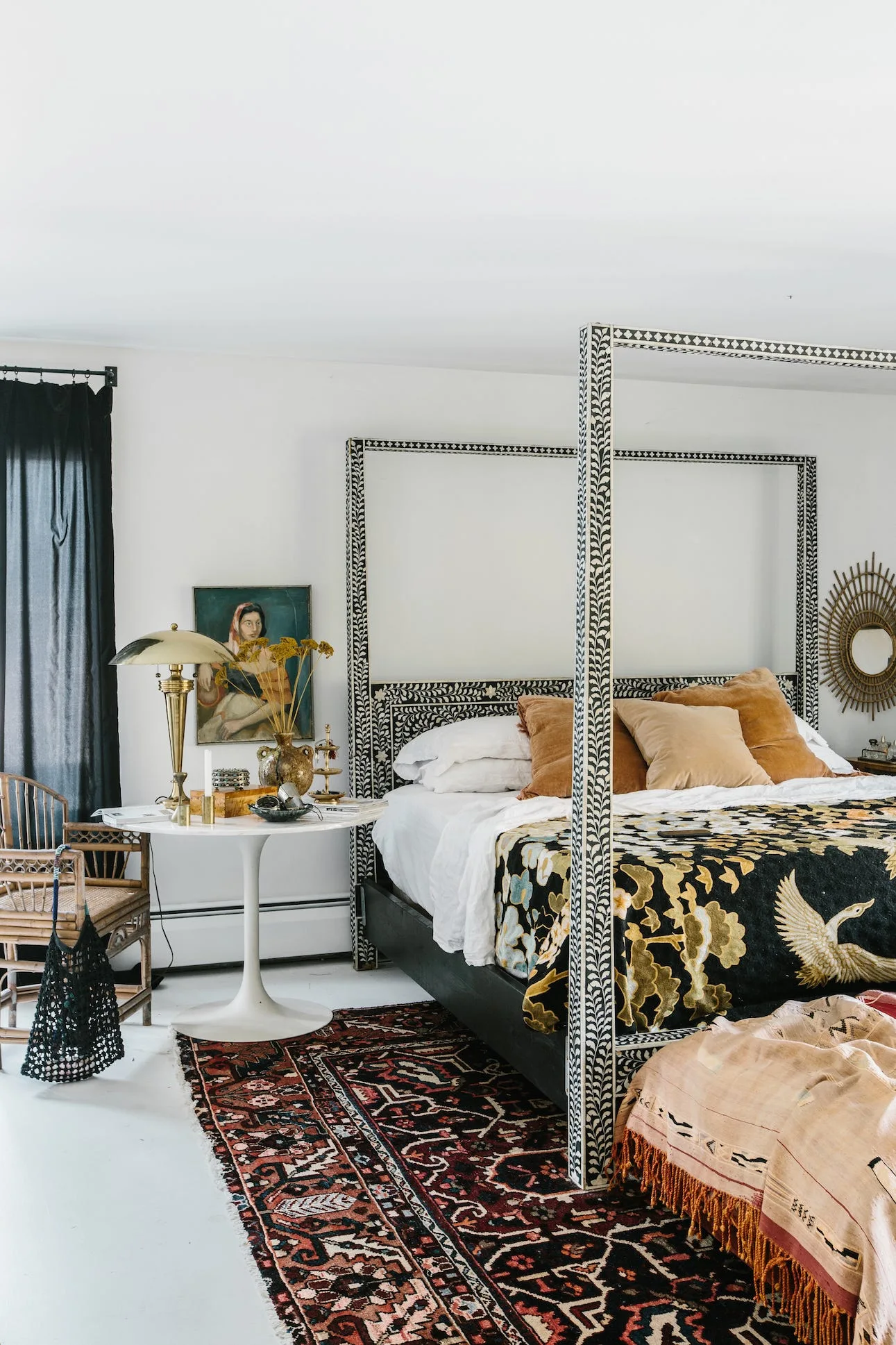 An eclectic bedroom features an intricate black and white patterned canopy bed, a white tulip nightstand with a brass lamp, and a dark oriental rug.