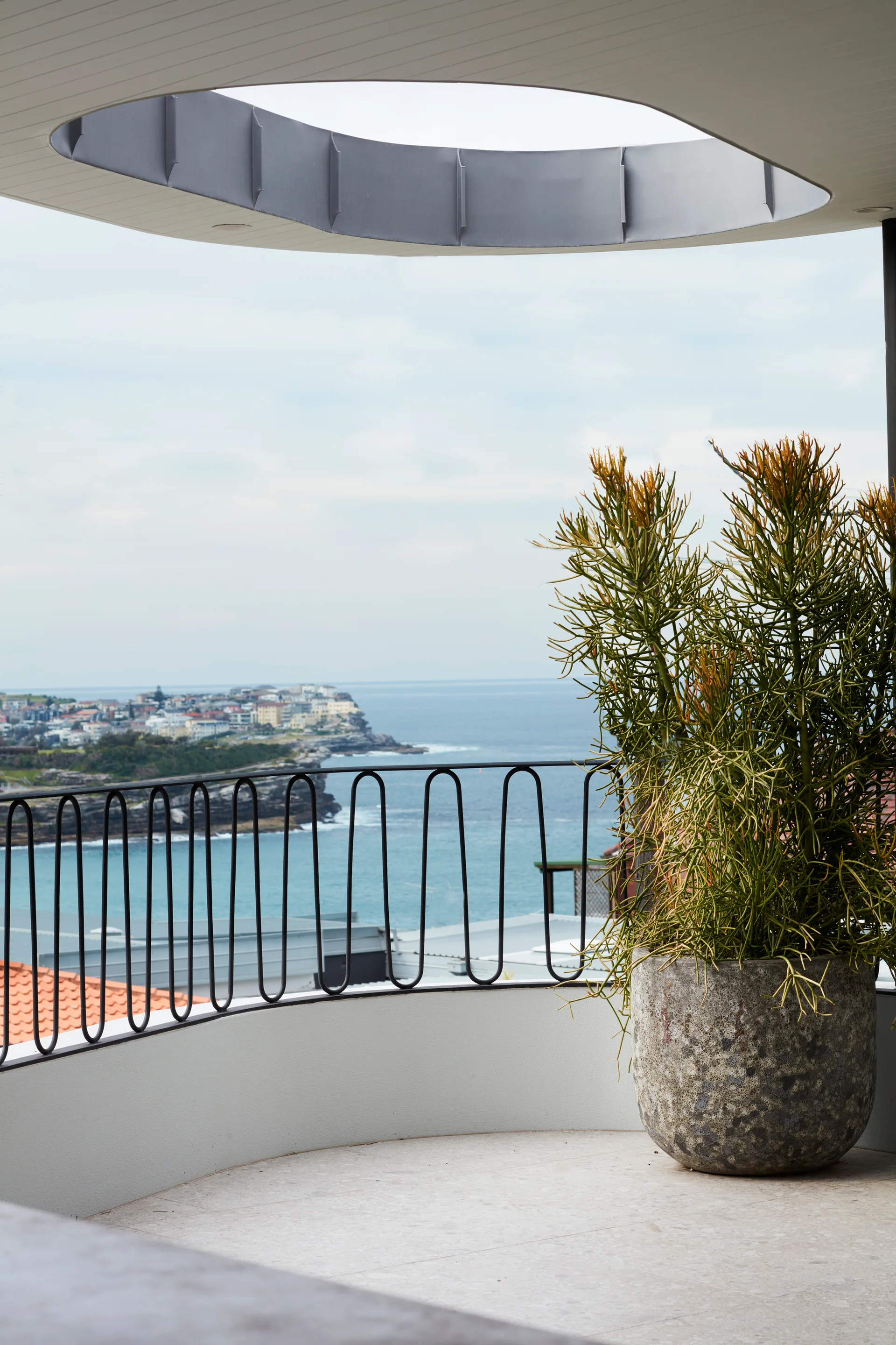 A curved balcony features a textured stone planter with a large plant, a wavy black metal railing, and an overhead ceiling cutout overlooking the ocean.