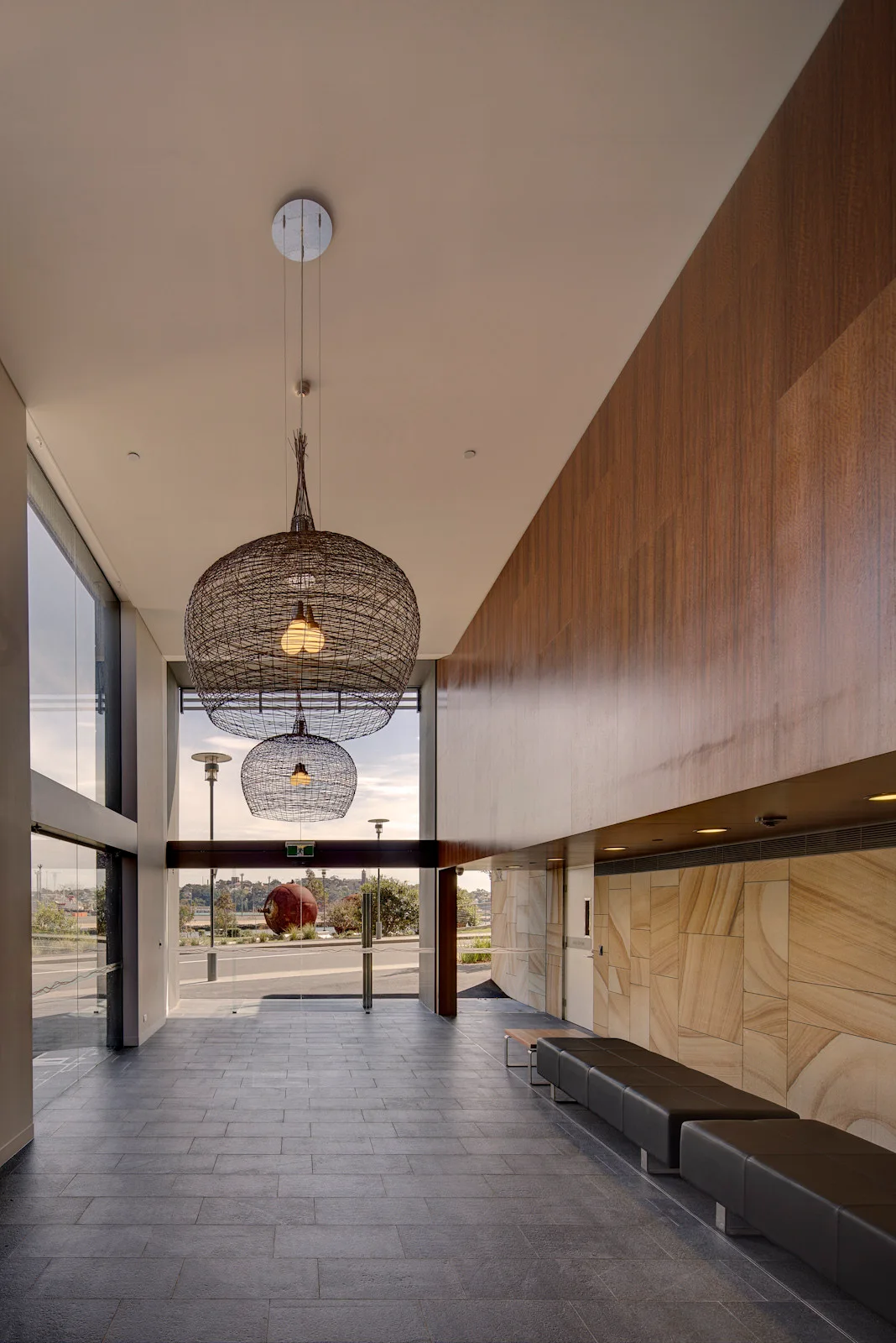 A spacious modern lobby featuring large wire mesh pendant lights, a sandstone wall, wood paneling, and black leather benches.