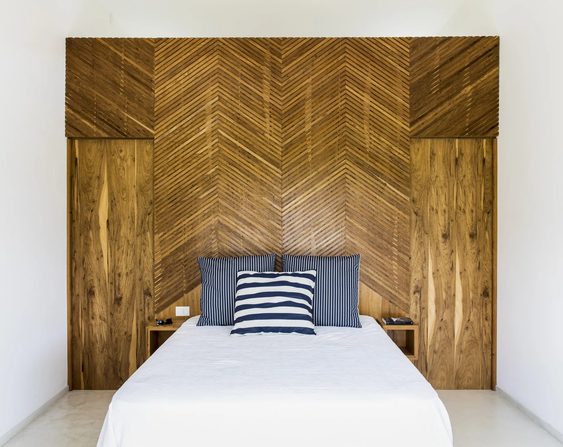 A bed with blue striped pillows is set against a full-height wood paneled wall featuring a central herringbone pattern and built-in cubby nightstands.
