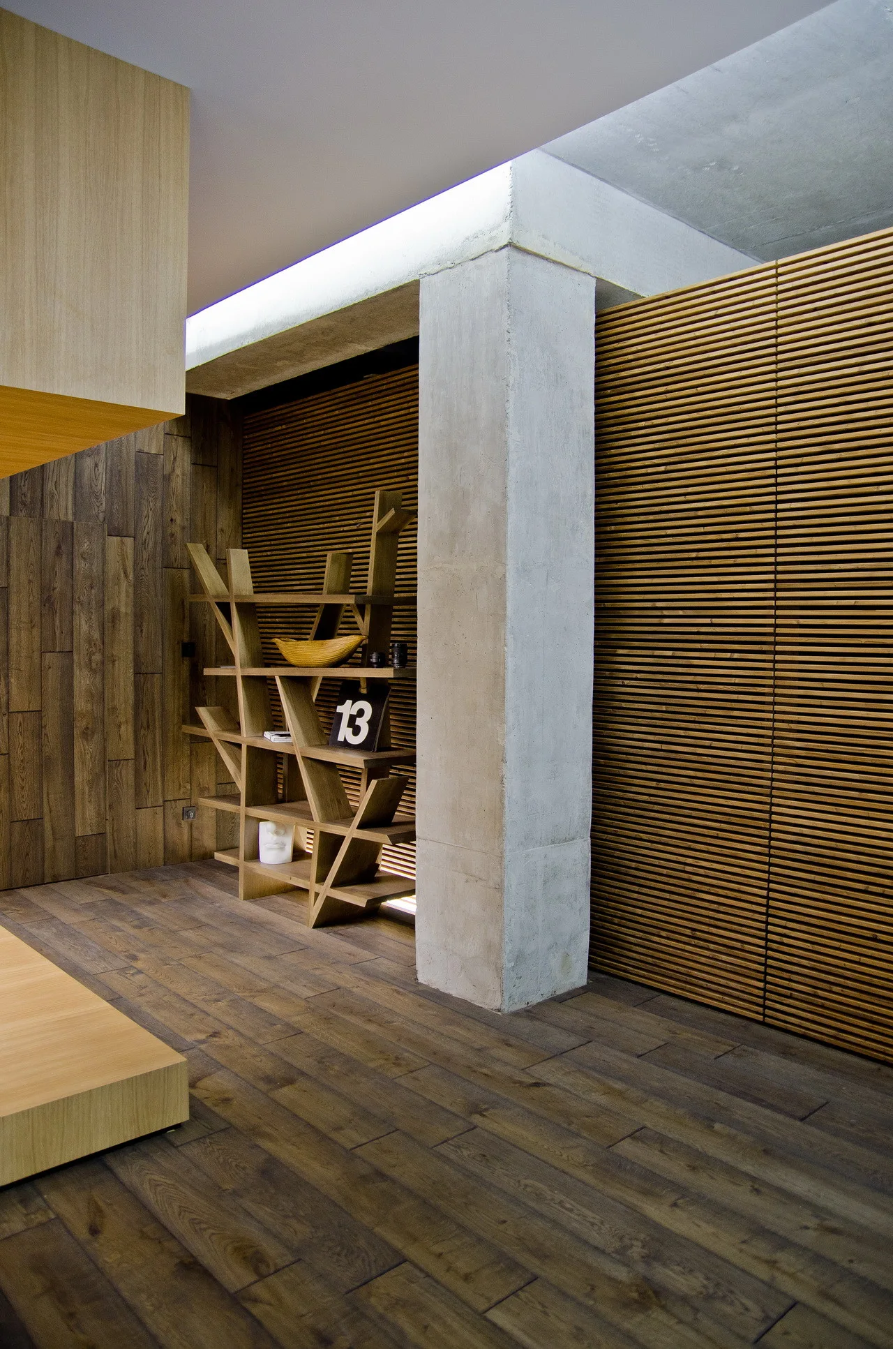 An interior space features horizontal slatted wood walls, a custom wooden bookshelf, a dark wood plank floor, and a raw concrete structural column.