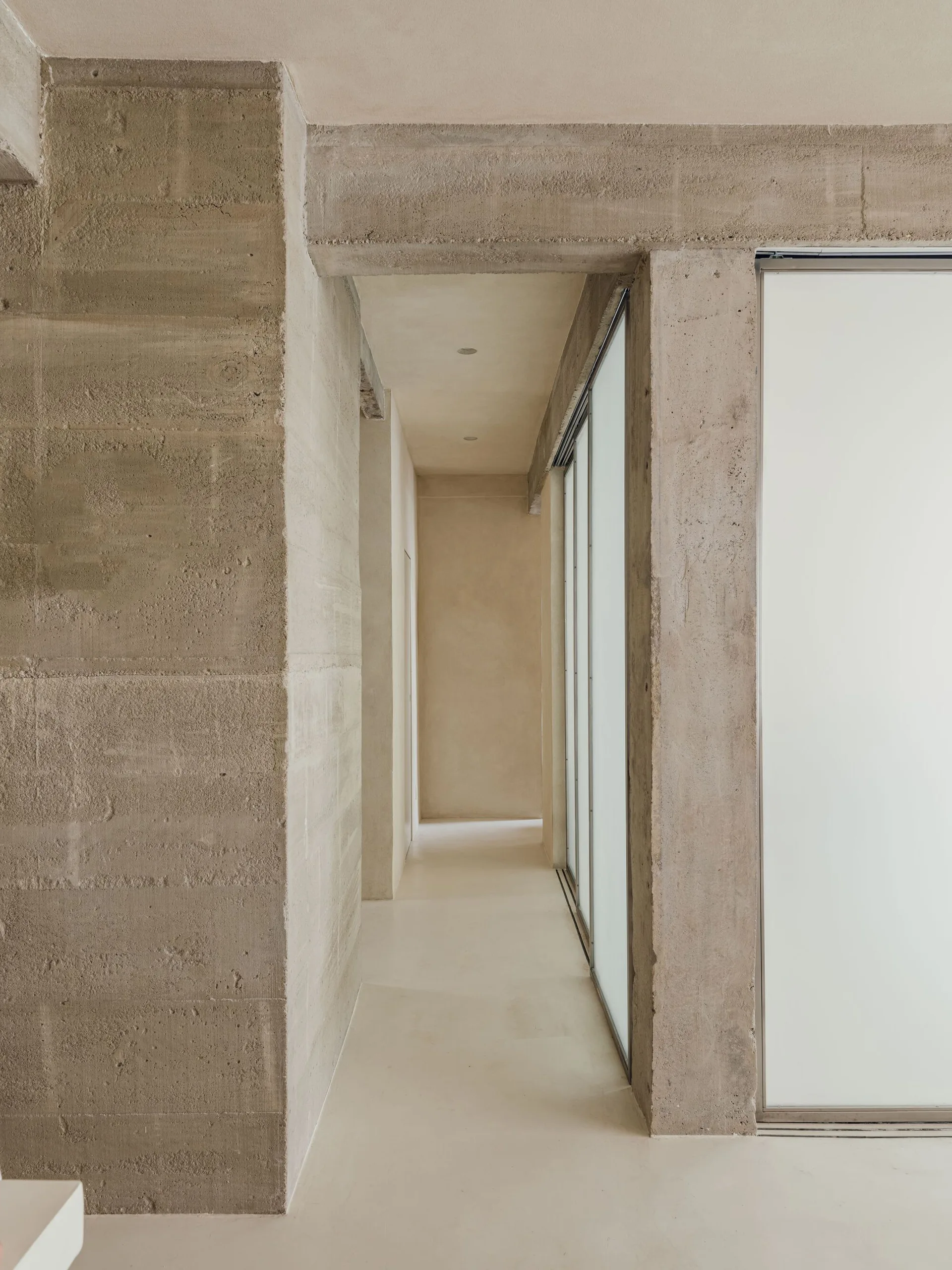 A minimalist hallway features textured raw concrete columns, smooth beige plastered walls, and large frosted glass sliding panels.