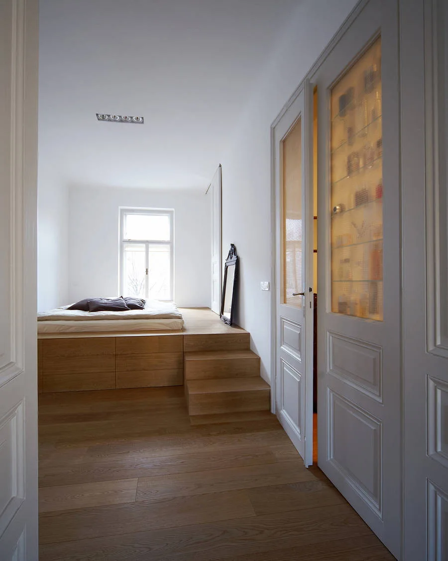 A minimalist white bedroom featuring a raised wooden platform bed with steps and built-in storage drawers beneath a central window.