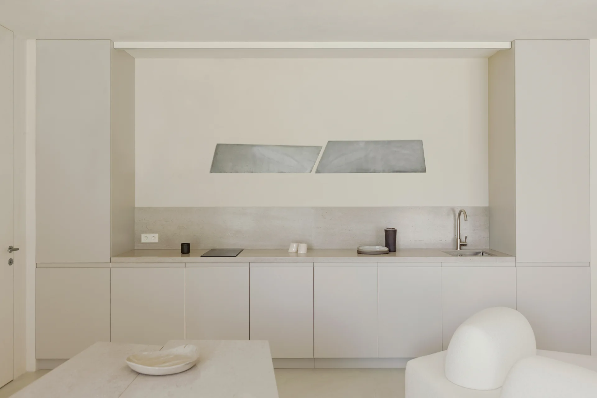 A minimalist beige kitchen features recessed flat-front cabinetry, a matching stone countertop with a single faucet, and a modern gray art piece.