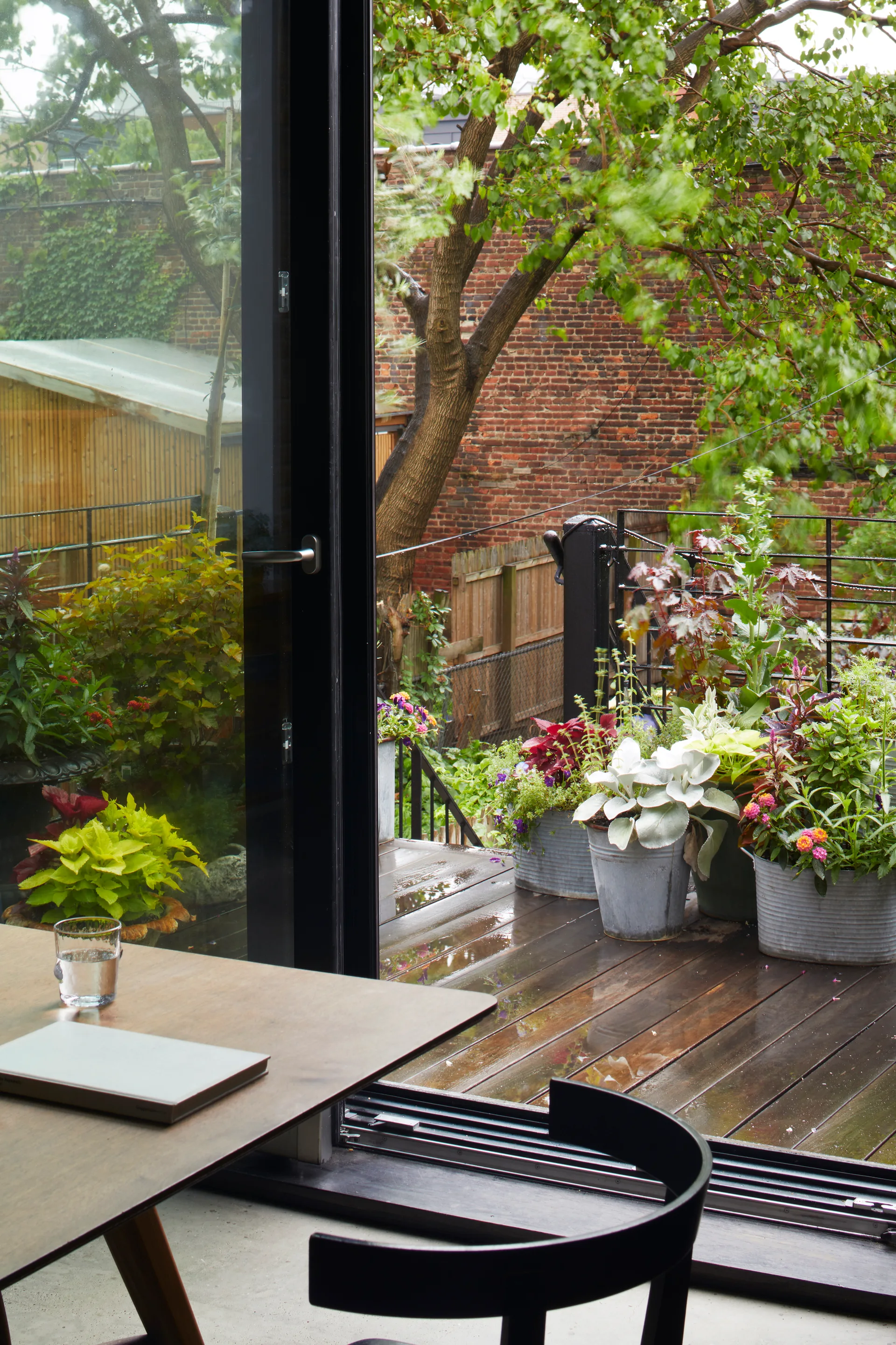 Looking from an indoor dining table through a large black-framed sliding glass door onto a wet wooden balcony filled with potted plants.