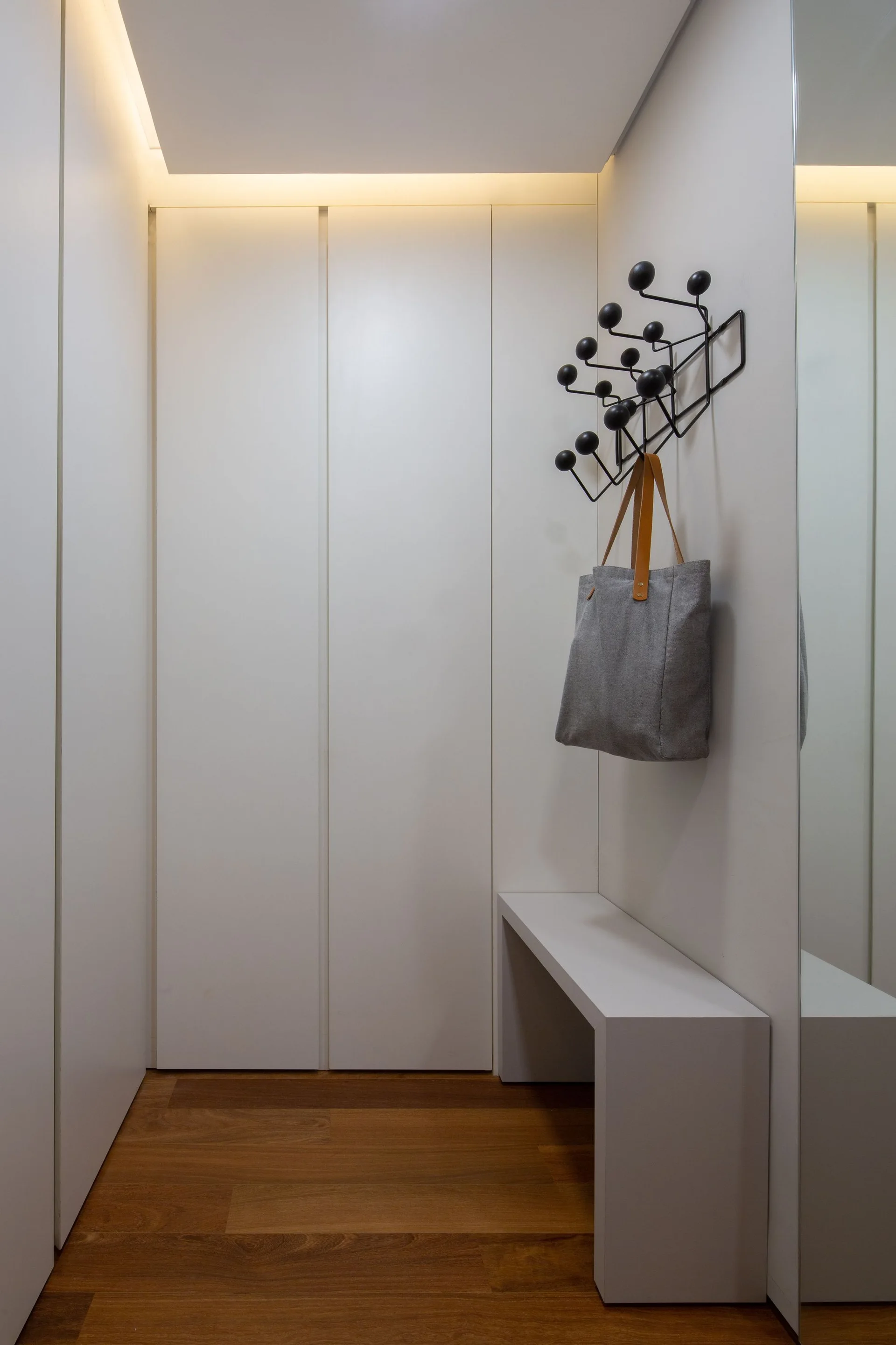 A minimalist white entryway featuring floor-to-ceiling handleless cabinets, a low matching bench, a black wall-mounted coat rack, and warm wood flooring.