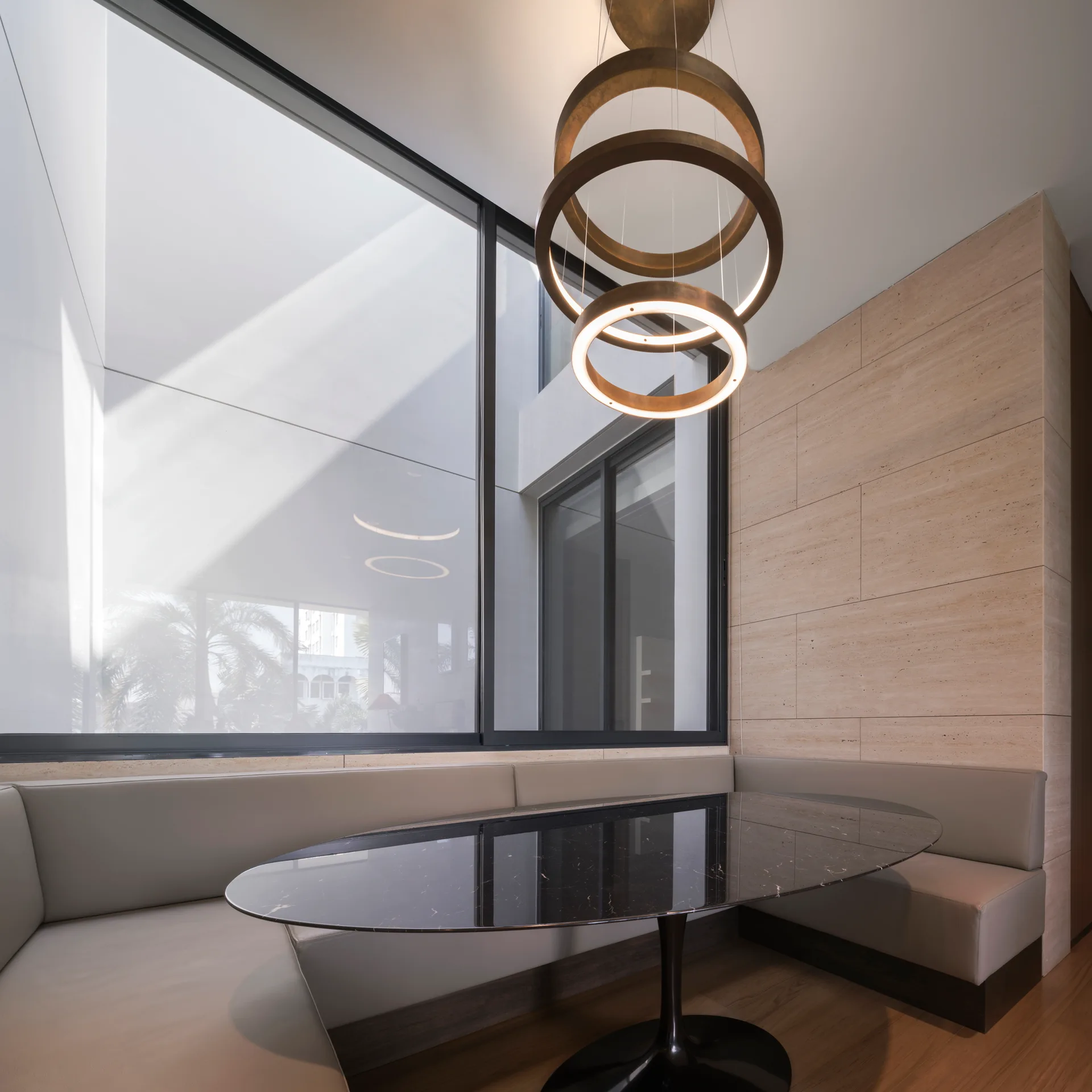 A modern dining area features a U-shaped gray banquette, a black oval marble table, a stone-clad wall, and a multi-ring pendant light.