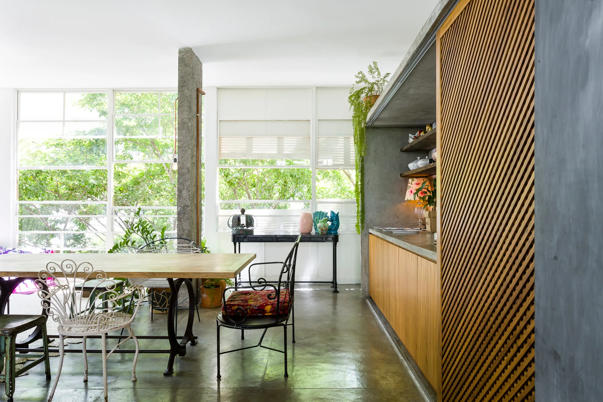 A light-filled dining space displays a wooden table, eclectic metal chairs, a polished concrete floor, and a sliding diagonal wood-slat screen.