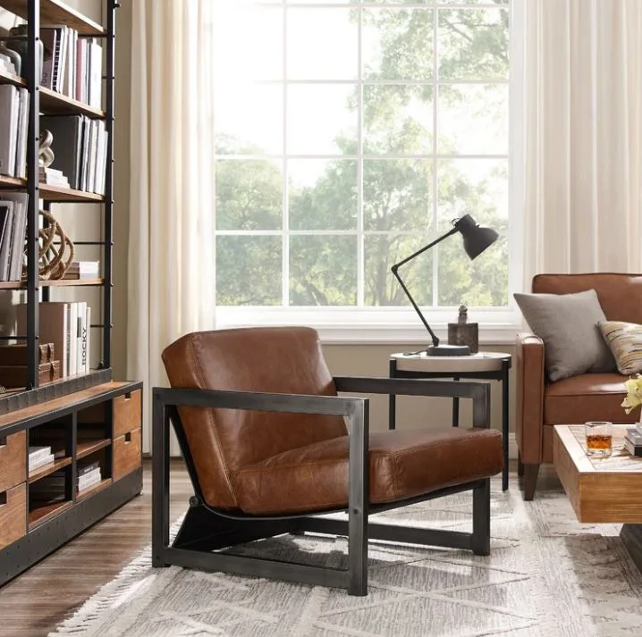A brown leather armchair with a black metal frame sits on a patterned rug next to an industrial bookshelf and a large window.