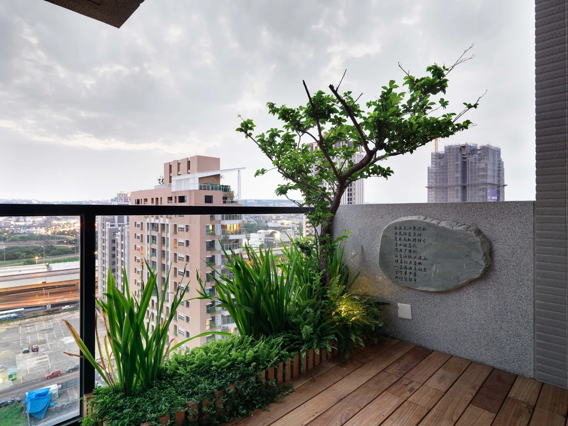 A high-rise balcony with wooden deck floors, a leafy built-in planter, and a stone calligraphy plaque on a grey textured wall.