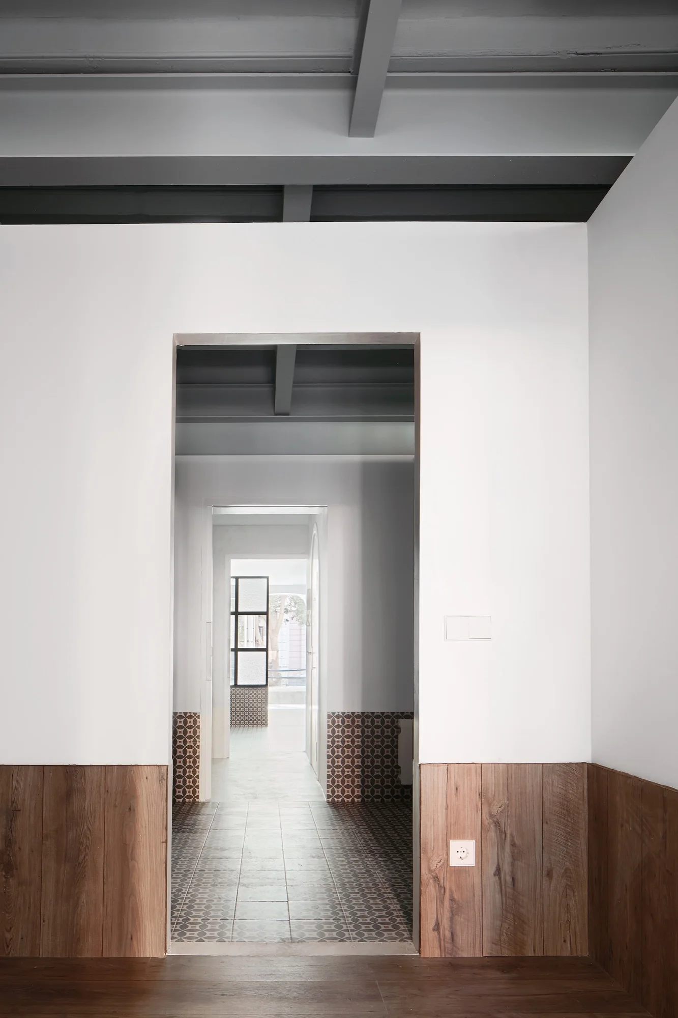 An interior hallway view looking through sequential doorways, featuring wood wainscoting, patterned tile floors, and grey exposed ceiling beams.