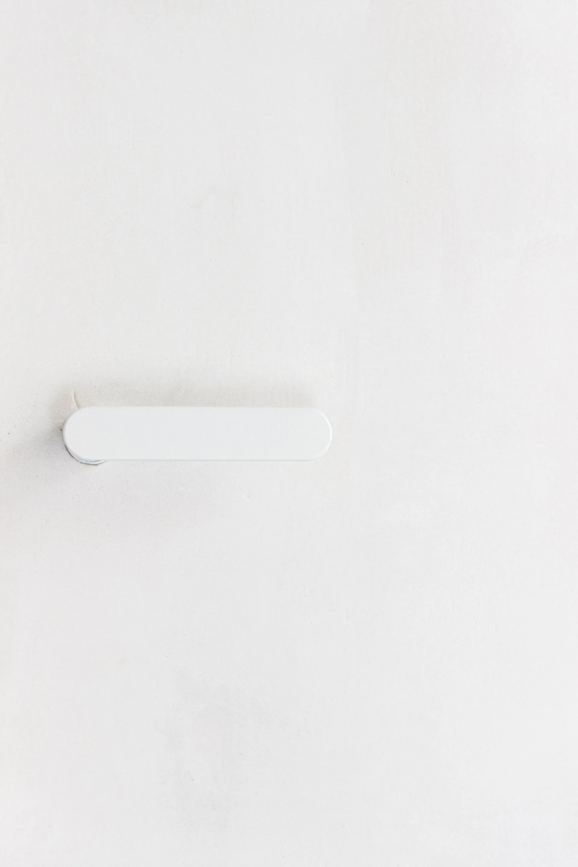 A close-up view of a minimalist matte white door handle with rounded edges mounted flat against a smooth matching white surface.