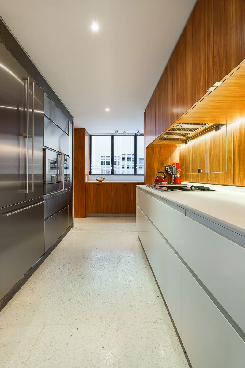 A galley kitchen featuring wood-toned upper cabinets, white base cabinets, integrated stainless steel appliances, and light terrazzo flooring.