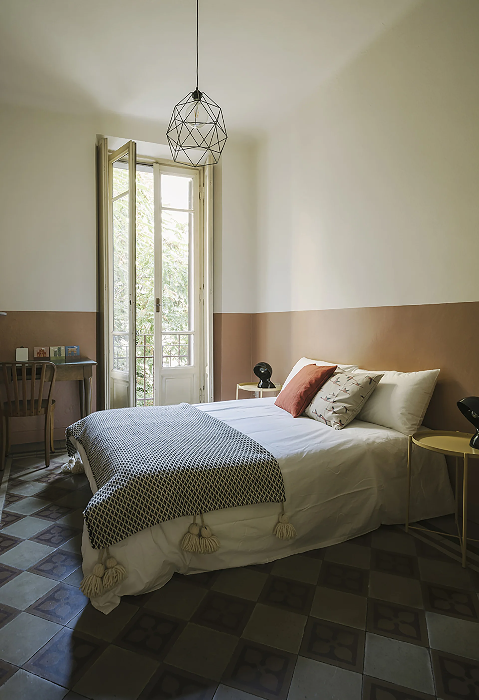 A bedroom features white and tan two-tone walls, patterned tile floors, and a bed with white sheets and a tasseled throw near open French doors.